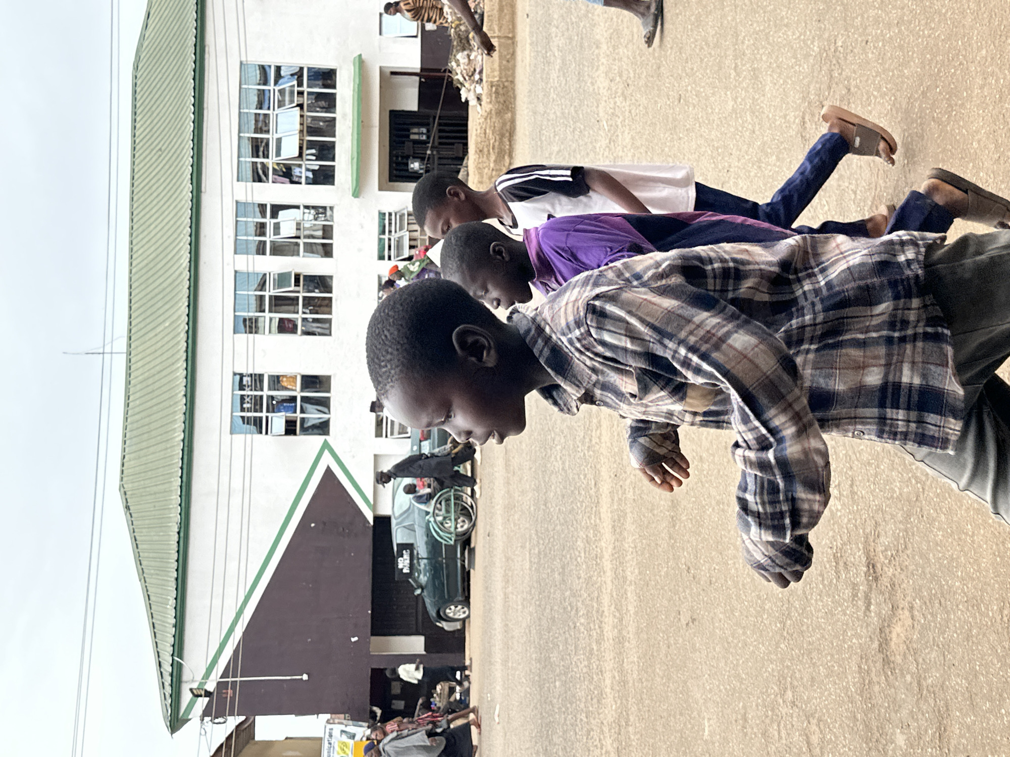 In Bukuru, Nigeria, a group of boys walks along a dusty, paved road. The foreground features a young boy with dark skin, wearing a plaid shirt and dark pants, walking towards the right side of the frame with his mouth slightly open, looking to his left. Behind him, another boy in a purple shirt and dark pants, followed by a boy in a white shirt with black stripes and dark pants, also walk to the right. In the middle ground, a dark green car is parked on the side of the road, with a sign that reads "NO PUBLIC" visible on a stand. Further in the background, a two-story building with a green metal roof and white exterior walls is visible. The building has numerous windows, some of which are open, and clothes appear to be hanging inside or outside some of them. The street is lined with various people and objects, suggesting a bustling marketplace or residential area. The sky is overcast.
