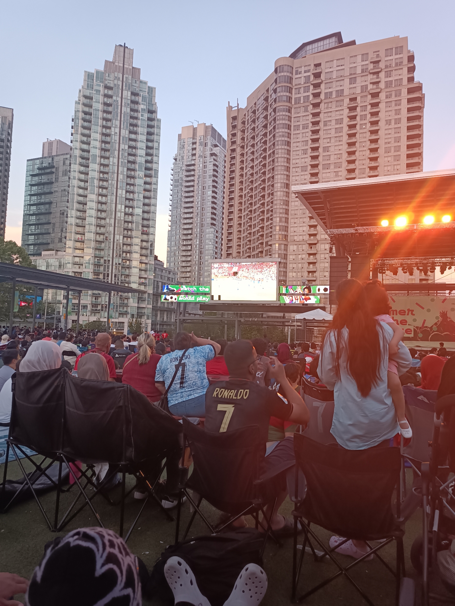 This image captures an outdoor gathering in Mississauga, Canada, likely at Celebration Square, where a crowd is watching a large screen displaying a soccer game. Several high-rise buildings loom in the background, characteristic of the city's urban landscape. People are seated in camping chairs and on blankets on a green artificial turf surface, facing a stage with bright lights. A large screen shows live sports action, with some text overlayed that reads "Watch the World Play." A woman in the foreground is holding a young child. The overall atmosphere suggests a community event, possibly a viewing party for a major sporting event like the FIFA World Cup. The sky is a soft orange and pink hue, indicating it is either sunrise or sunset.