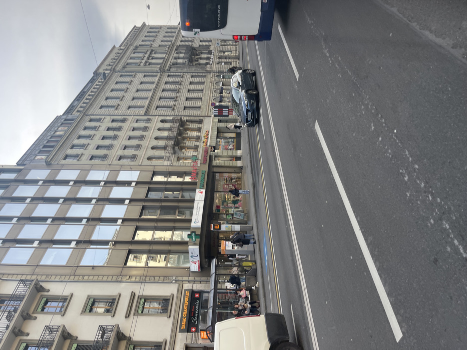 This media file captures a street scene in Geneva, Switzerland, under an overcast sky during daytime. The image is oriented vertically but depicts a horizontal street view.
On the right, an asphalt road features white lane markings, with a partial view of the rear of a white and blue bus, bearing the name "GITARD." Further down the road, a dark-colored car is visible, possibly parked or moving slowly.
The left side of the scene is dominated by urban architecture. A tall, multi-story building displays classical European design with numerous windows, decorative cornices, and ornate balconies on its upper floors. At street level, various shops occupy the ground floor. Visible signage includes "PHARMACIE" (French for Pharmacy) and "Parapharmacie," indicating a pharmacy. Another prominent sign reads "TABAC CHANTEPOULET" (Tabac referring to a tobacco shop) and "Souvenirs," distinctly featuring the Swiss cross emblem, which helps confirm the Geneva location, as "Chantepoulet" is a street in the city. Adjacent to this, a more modern building with a facade of large, reflective glass windows is also visible.
Several pedestrians are present on the sidewalk. Some individuals are seen near the shop entrances, while others are walking or waiting near what appears to be a bus stop area. One person is positioned in the street near the dark car. The overall setting depicts a typical busy urban environment with a blend of historical and contemporary buildings.