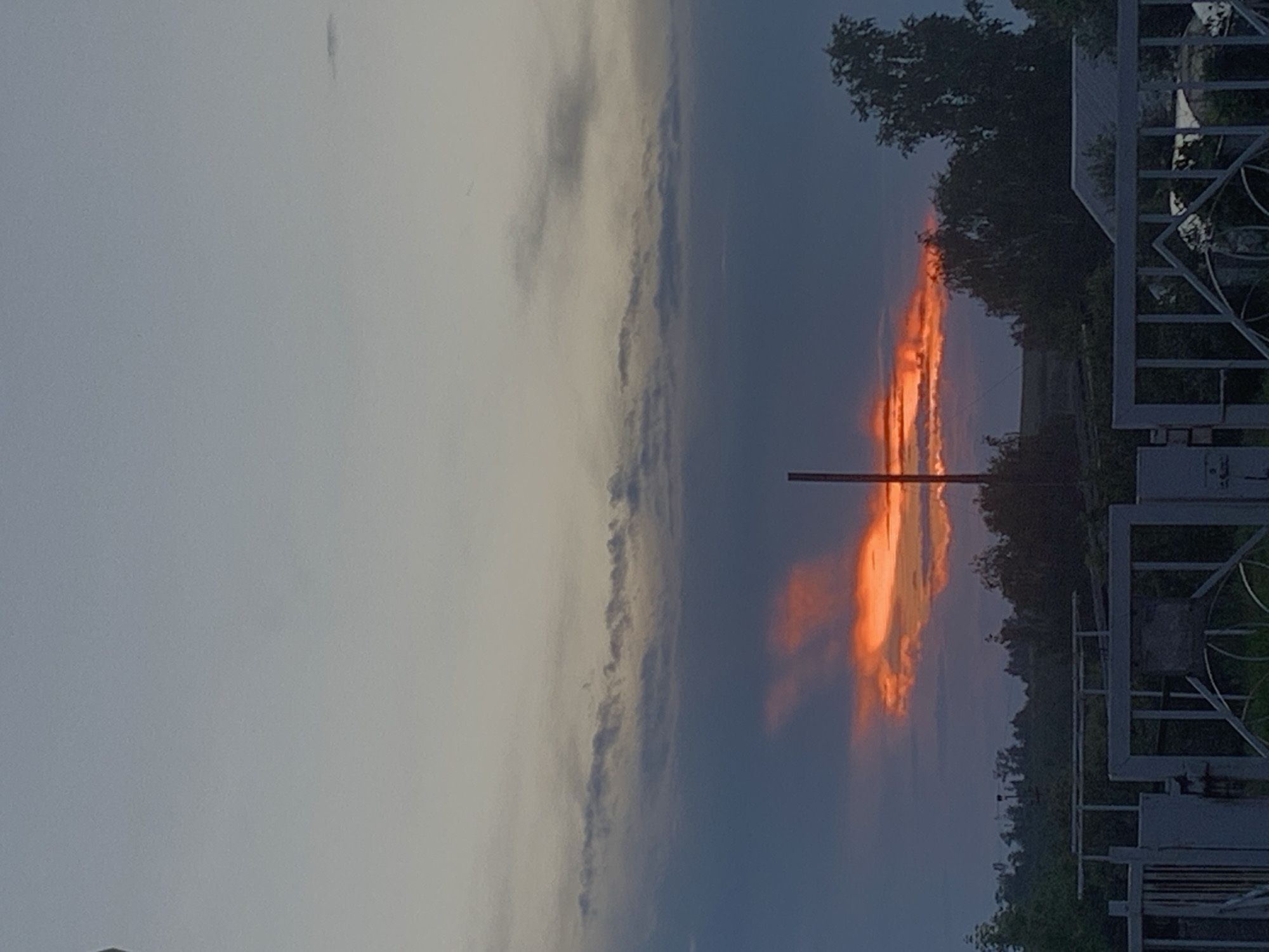 The image depicts a rural or suburban landscape at dusk. The sky is predominantly a gradient of pale blue and grey, with darker clouds obscuring the upper portion. A dramatic band of vibrant orange and red clouds is visible towards the horizon, suggesting the setting sun is peeking through or illuminating the undersides of the clouds. The foreground features a metal fence with decorative elements and a gate. Beyond the fence, there is a dense line of trees and vegetation, with some scattered buildings, possibly houses or outbuildings, partially visible amongst the foliage. The overall impression is of a peaceful, natural scene during twilight, with a striking display of natural light. The location is identified as Фоки, Russia.
