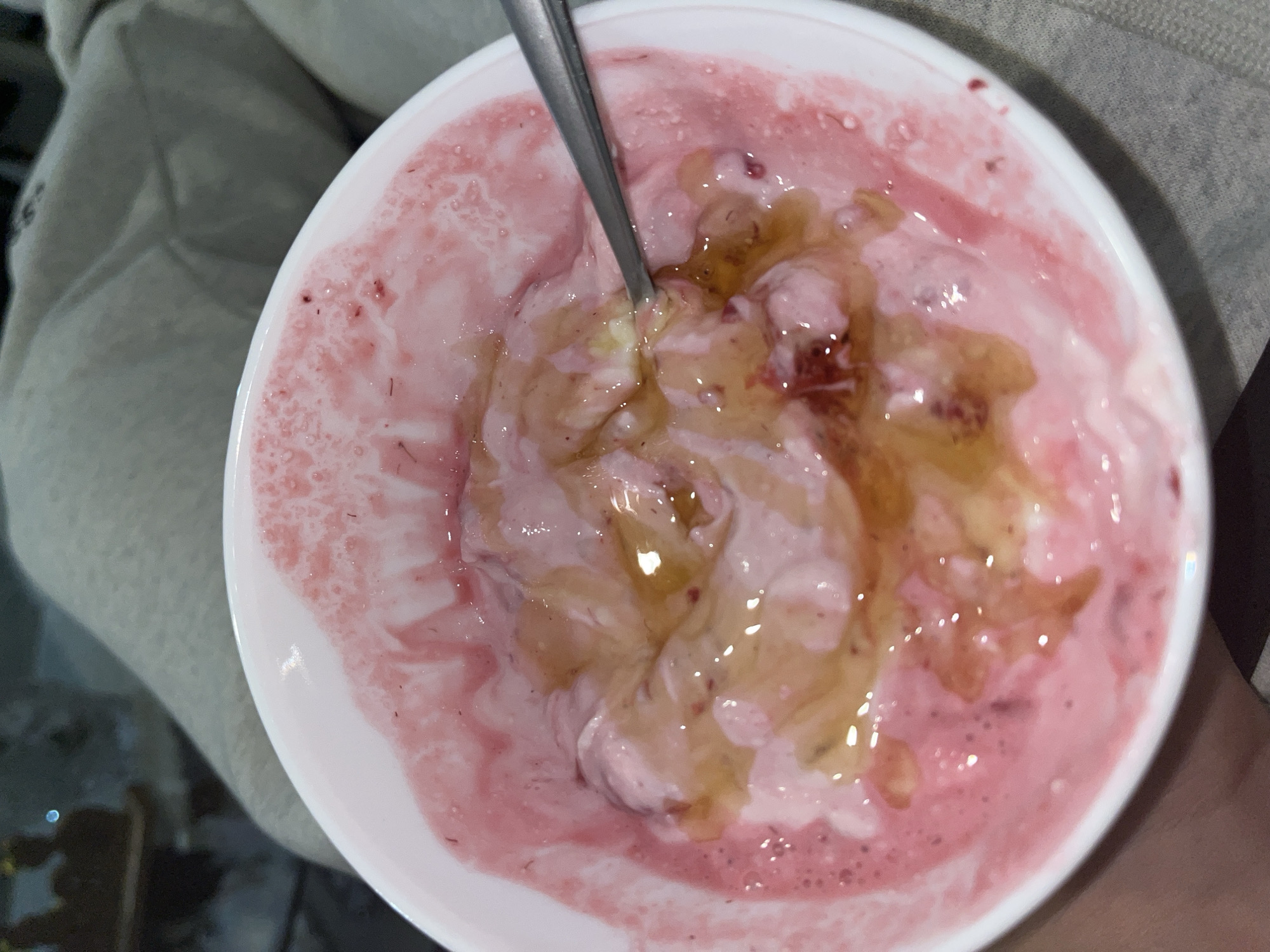 A close-up, top-down view shows a white bowl filled with pink yogurt and drizzled with honey. A silver spoon rests inside the bowl. The yogurt appears to have been partially eaten, with swirls of pink and white visible. The honey creates glossy patches atop the yogurt. The bowl sits on a surface covered with light-colored fabric. No people are visible. The location is unknown.