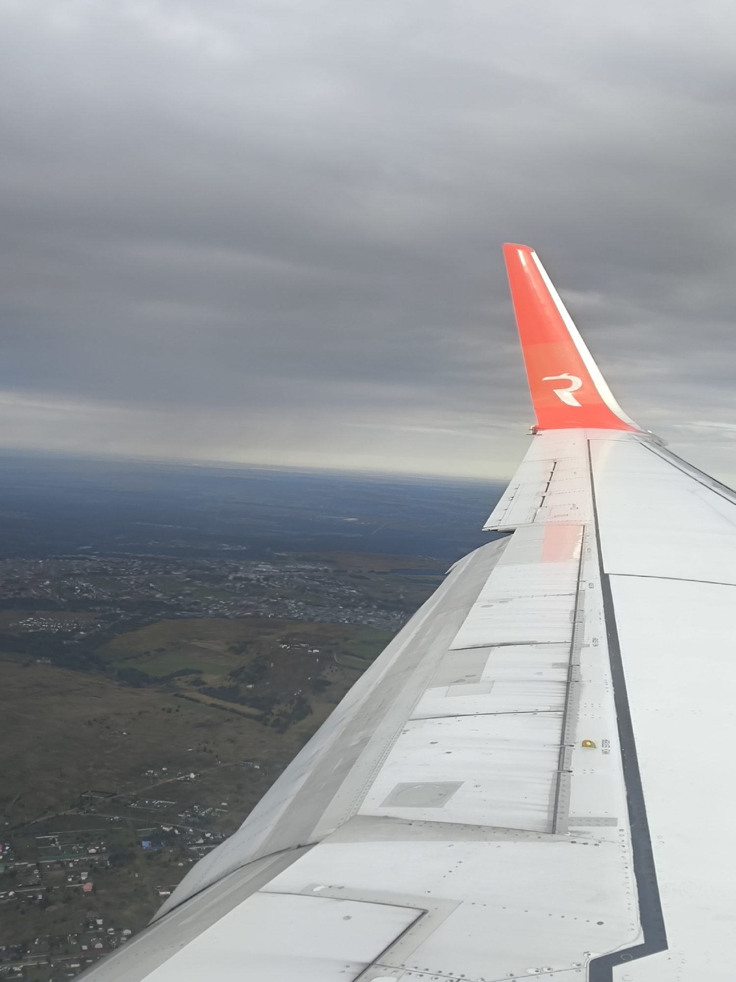 An aerial view from an airplane shows a wing with a red tip featuring a white stylized "R" logo. Below the wing, a landscape unfolds with houses, roads, and greenery, suggesting a rural or suburban area. The sky is overcast with gray clouds.