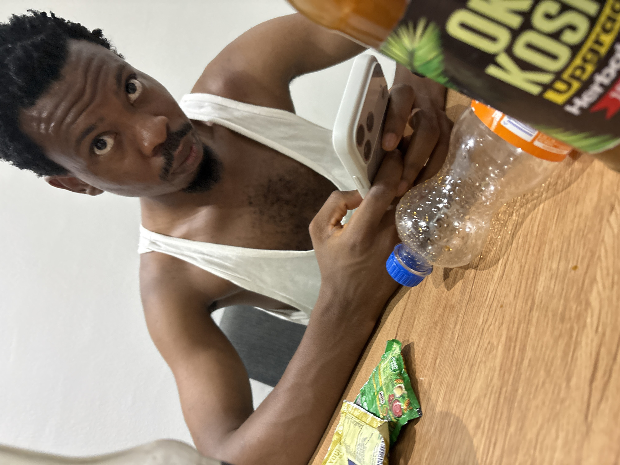 The image features a young adult Black man looking directly at the camera with a focused, intense gaze. He has short, dark, curly hair, a mustache, and a goatee, and is wearing a white tank top, which reveals some chest hair. The photo is tilted at an angle, giving it a dynamic composition.
The man is holding a white-cased smartphone, which has a visible triple camera setup on its back. His hands appear to be actively engaged with the phone. He is seated or leaning against a light-colored wooden table that occupies the foreground and right side of the frame. The background is a plain white wall, suggesting an indoor setting.
On the table, several items are visible. In the upper right corner, a large bottle, partially obscured, contains a brown liquid. Text on this bottle includes "OK KOS...", "UPGRAd...", and "Herbal". Beside it is a clear, empty plastic bottle with a bright blue screw cap. Further down on the table, two small sachets are present: one green, partially revealing "CHOCO..." text, and another yellow sachet.
The lighting appears bright and even, consistent with an indoor setting during daytime or under strong artificial light. The man's direct eye contact and serious expression are prominent emotional elements.