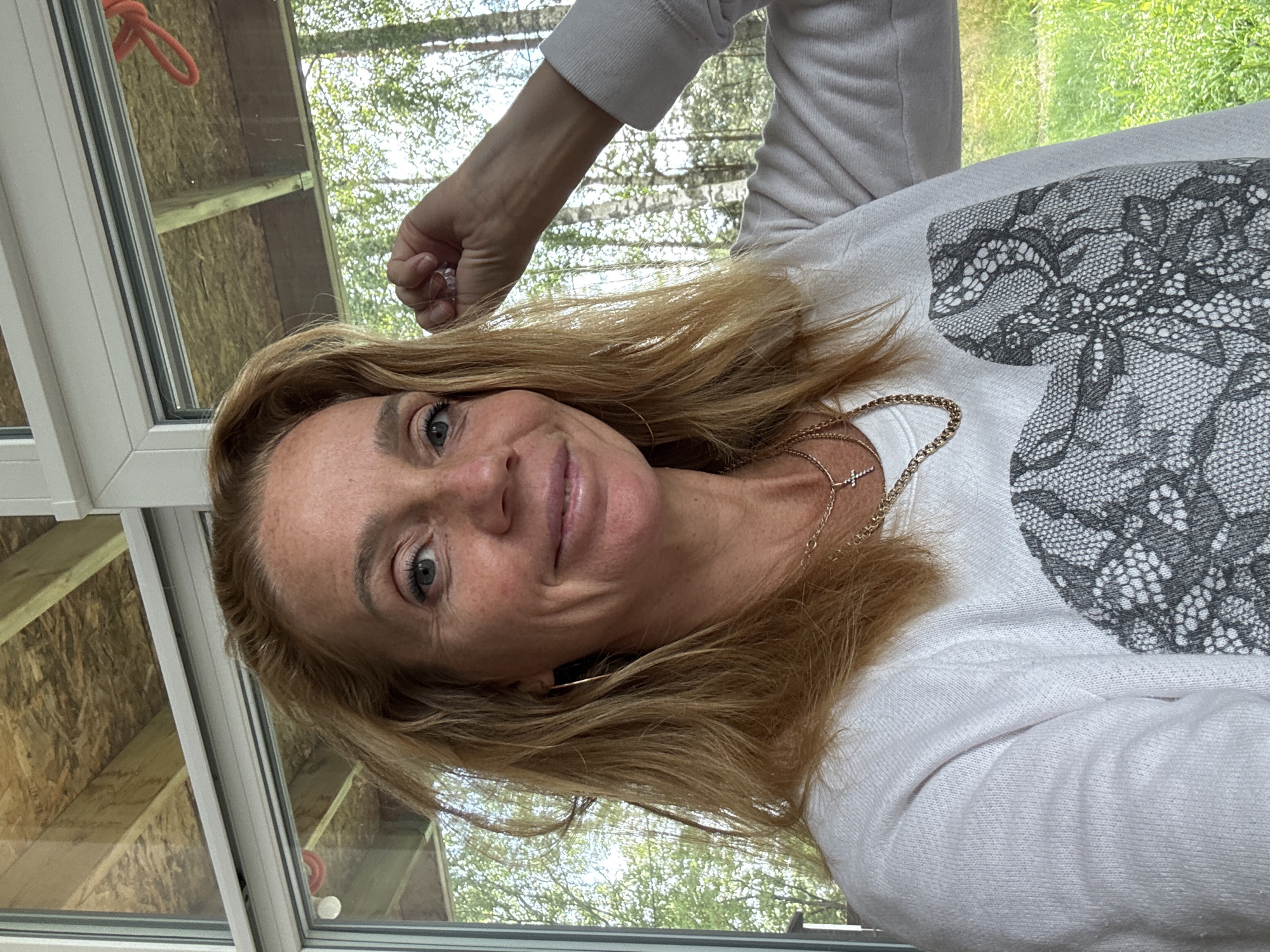 A candid, medium shot captures a woman with shoulder-length, wavy, blonde hair and fair skin with freckles. She is looking directly at the camera with a slight smile. Her right arm is raised, with her fist clenched near her head. She is wearing a white sweatshirt with a black lace design on the front, and a gold chain necklace with a cross pendant. The background shows a large window with a view of trees and greenery, suggesting an outdoor or semi-outdoor setting, possibly a porch or a room with a large window. The ceiling structure with exposed beams and OSB sheeting is visible above the window. The location is described as Steklyannyi, Russia.