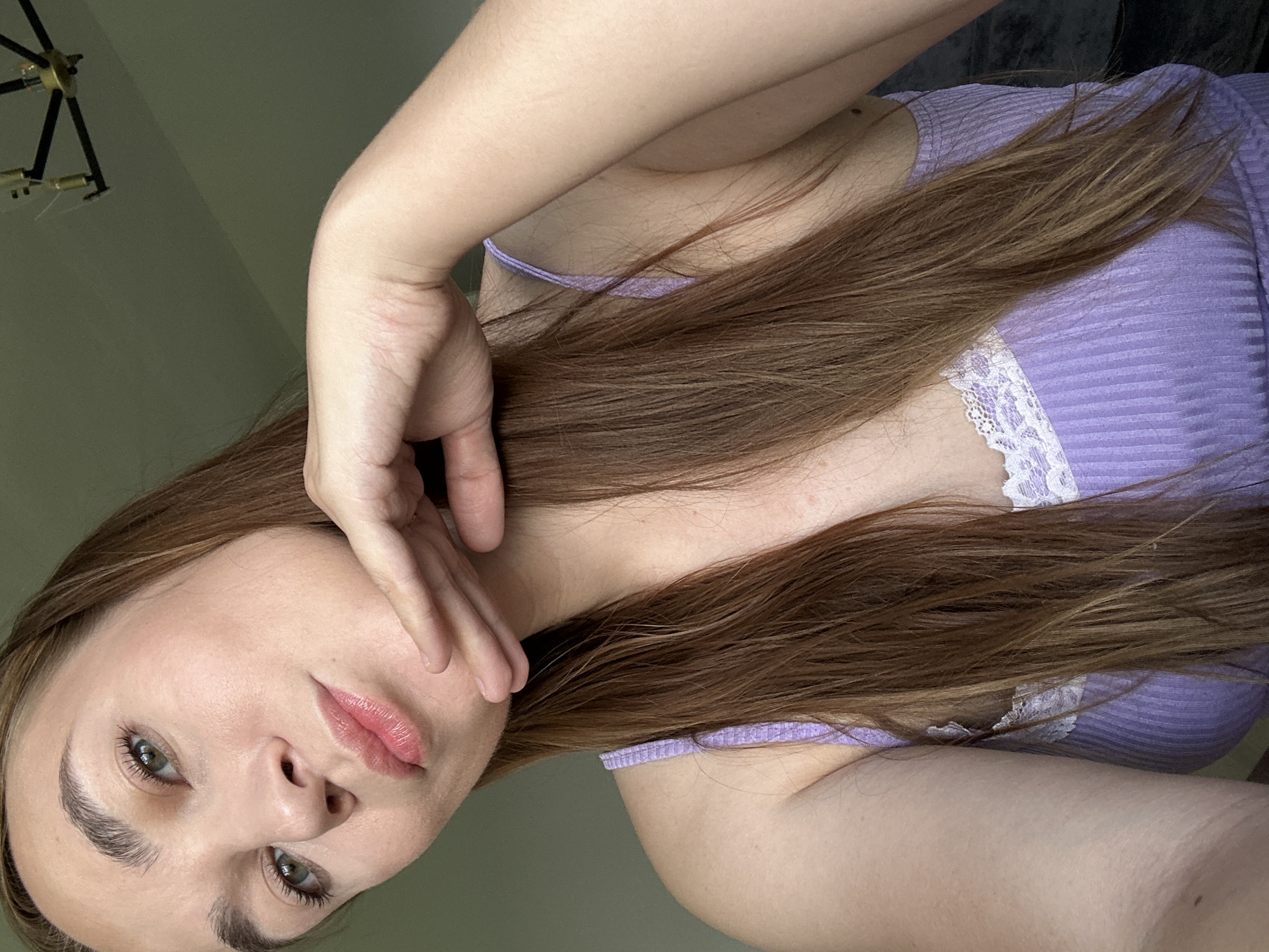 This is a close-up, vertical, selfie-style photo of a person's face and upper body. The person has long, reddish-brown hair that falls over their shoulder and chest. They are wearing a ribbed, lavender-colored tank top with white lace trim. Their skin appears fair and smooth, and they have green eyes. Their left hand is raised and gently touches their cheek and mouth area. Their lips are a soft pink. The background is a plain, muted green wall. A portion of a black and gold chandelier-style light fixture is visible in the upper left corner. The lighting suggests it could be daytime indoors. The overall mood of the image is calm and introspective. There is no visible text in the image.