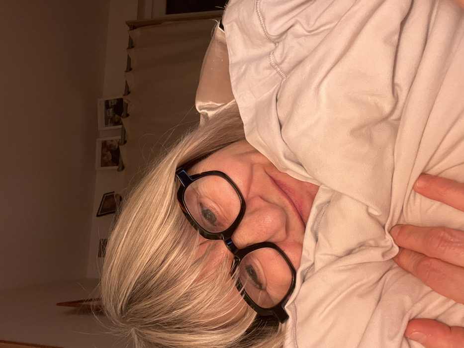 An indoor, dimly lit, close-up shot captures a person, likely in a bedroom setting in Chambœuf, France. The subject, appearing to be an adult with light blonde or grey hair, is seen from the head to upper torso. They are partially obscured by a light-colored, soft blanket or duvet, which is pulled up around their face and neck, covering a significant portion of their head and body.
The person is wearing distinct black, thick-framed eyeglasses. One eye is clearly visible through the lens, looking directly at the viewer, revealing a green or blue iris. The nose and part of the mouth are also visible, showing natural facial creasing. A hand is seen holding the fabric of the blanket on the right side of the image.
The background, seen to the left of the person's head, consists of a soft, warm-toned wall. Several small, framed pictures are hanging vertically on the wall. Above these, a window with a rolled-up blind or curtain is partially visible. The overall lighting is soft and warm, suggesting either an early morning, late evening, or a dimly lit room. The person appears relaxed and cozy, perhaps just waking up or settling down in bed. The image is taken at a slight tilt, adding a dynamic angle to the composition. No visible text is present in the scene.