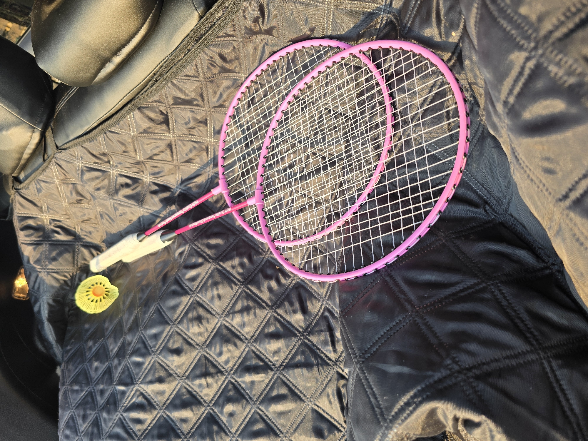Two pink badminton rackets and a yellow shuttlecock lie on a black quilted fabric surface. The fabric appears to be a car seat cover. The background consists of dark car seats and a reflection of light. The image is taken from a high angle, looking down at the objects. There are no people visible in the image.