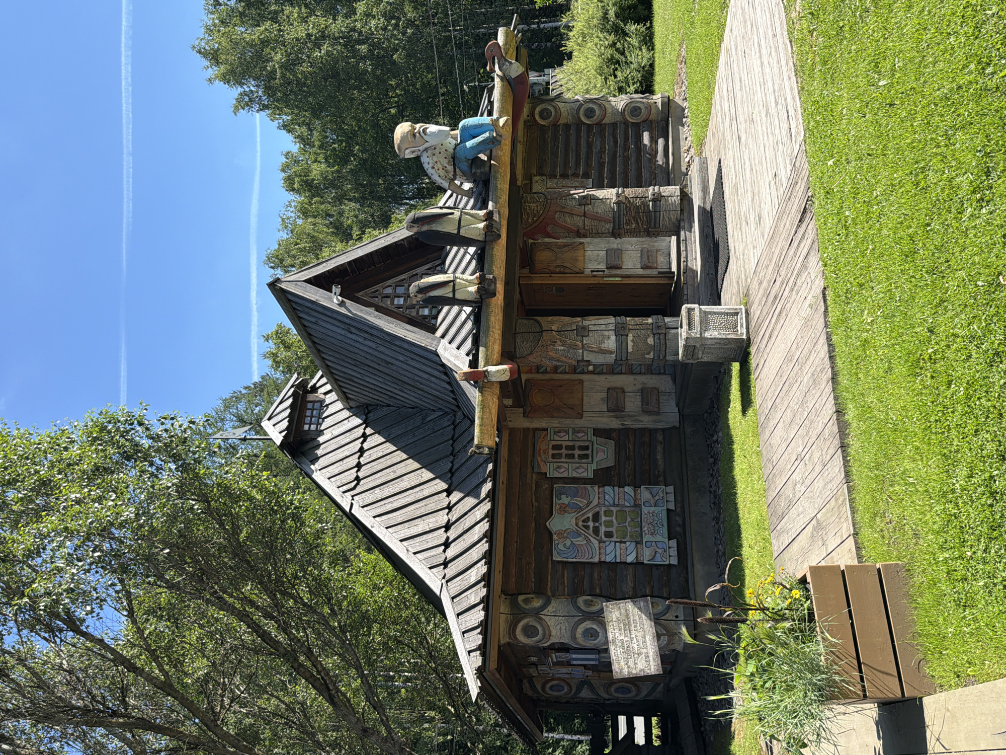 A medium shot captures a rustic wooden structure, possibly a small house or a museum exhibit, set against a backdrop of lush green trees and a clear blue sky with two faint contrails. The building features a steep, multi-gabled roof covered in wooden shingles, with a small dormer window on the front gable. The walls are constructed from large, horizontally stacked logs, and are adorned with intricate, colorful carvings and painted window frames. Above the entrance, a wooden beam is decorated with carved figures of birds and a seated old man with a white beard and blue pants. The structure is approached by a wooden deck platform that extends into a well-maintained grassy lawn. To the left of the entrance, a wooden sign with text is mounted on a post. In the foreground, a wooden planter box filled with tall green grasses and some yellow flowers sits on the left side of the deck. The scene is bathed in sunlight, casting shadows across the lawn and the building, suggesting a clear, bright day.