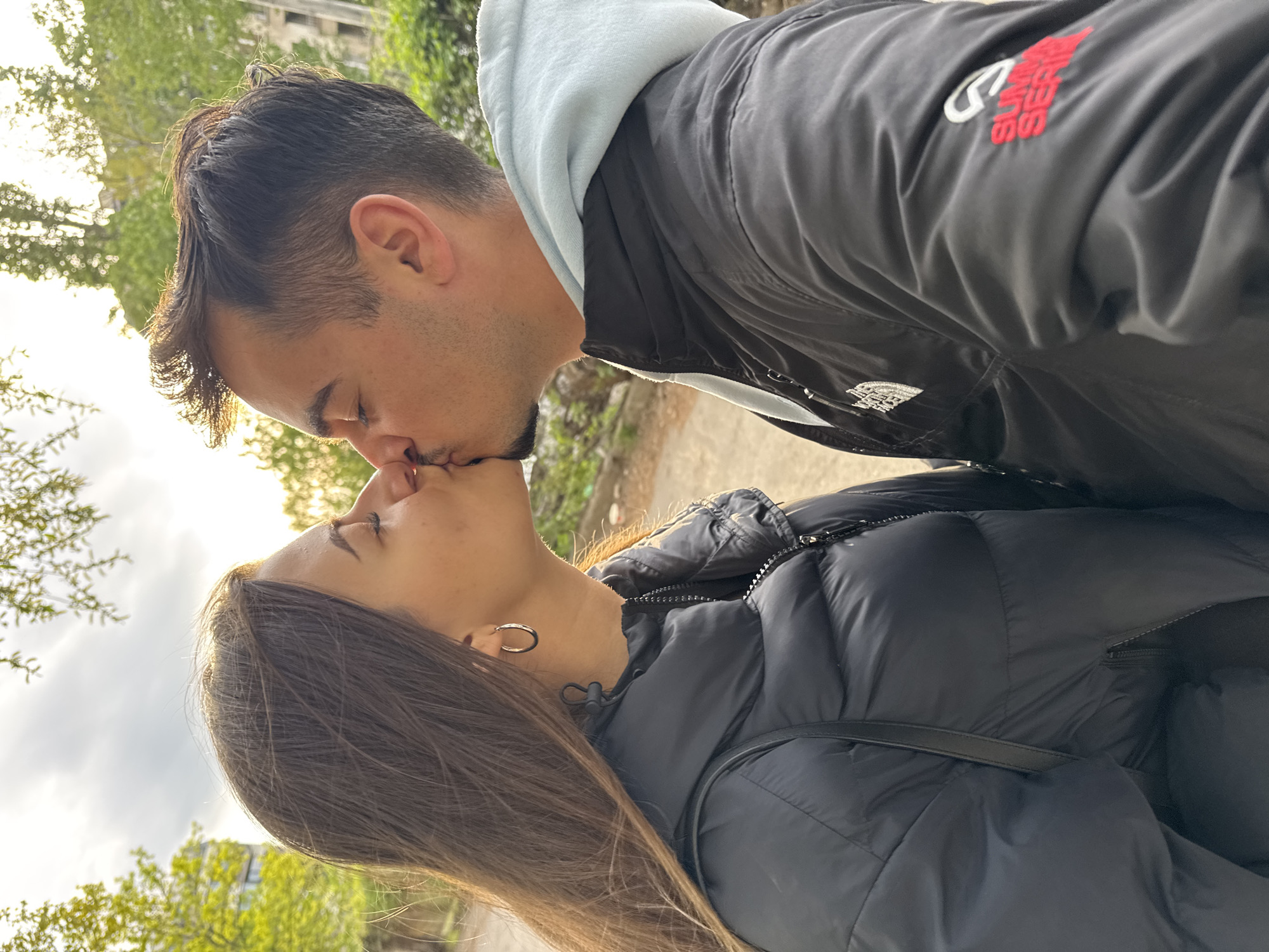 This is a vertically oriented, eye-level shot capturing a man and a woman sharing a kiss outdoors.
The main subjects are a man and a woman in an intimate embrace. The man, positioned on the upper right, has dark hair with lighter strands catching the sunlight, stubble on his face, and his eyes are closed. He is wearing a black jacket with a light blue hooded sweatshirt underneath. Visible on his black jacket are red letters spelling "SLUMBER" and possibly "SESSIONS" below it, along with a white, circular logo that appears to be a "C". Further down, a white emblem resembling "The North Face" logo is visible.
The woman, on the lower left, has long, straight brown hair, which is also illuminated by the sun. Her eyes are closed as she kisses the man. She is wearing a black puffer jacket and a silver hoop earring in her left ear. A black strap, possibly from a bag, is visible over her shoulder.
The activity taking place is a kiss, indicating affection and intimacy between the two individuals.
The setting is outdoors, likely in an urban or park environment, given the visible trees and sky. The background features bright, dappled sunlight filtering through green foliage, suggesting it is daytime, possibly morning or late afternoon. The sky is partially cloudy. While not explicitly visible in detail, the location context is provided as Sofia, Bulgaria. The overall mood is tender and romantic.
