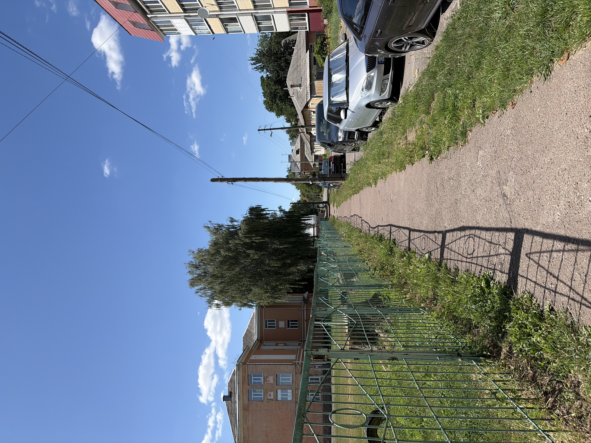 The photograph depicts a sunlit day in an urban environment, possibly in Eastern Europe given the architecture and vehicles. The scene is dominated by a paved pedestrian walkway on the right side, lined with grass and parked cars. A low green metal fence runs along the left side of the walkway, bordering a grassy area. Beyond the fence, an older, tan-colored building with multiple windows is visible, partially obscured by a large, mature tree with weeping branches. In the background, several other buildings, including a taller apartment building with a red-tiled roof, and smaller residential structures are visible. Power lines crisscross the bright blue sky, which is dotted with a few white clouds. The cars parked along the walkway include a silver hatchback, a dark SUV, and a dark sedan. The overall impression is one of a quiet residential street on a clear summer day.