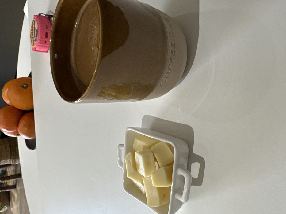A brown mug with "COFFEE TIME" inscribed is adjacent to a small, square white dish filled with cheese cubes. Oranges are clustered to the side of the mug, along with a pink measuring tape. All items rest on a white surface.