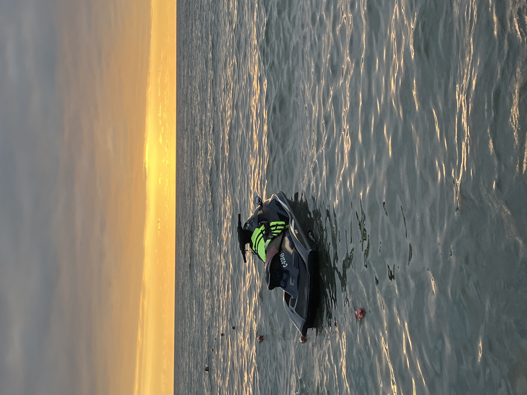 A black jet ski with a person wearing a neon yellow life jacket is floating on the water. The jet ski has the identification number "C-02645" on its rear. The water is calm with gentle waves reflecting the orange and yellow hues of the setting sun. Several small buoys are floating in a line to the left of the jet ski. The horizon is visible with a band of orange and yellow light from the sun peeking through gray clouds. The scene suggests an outdoor recreational setting, likely a beach or coastline.
