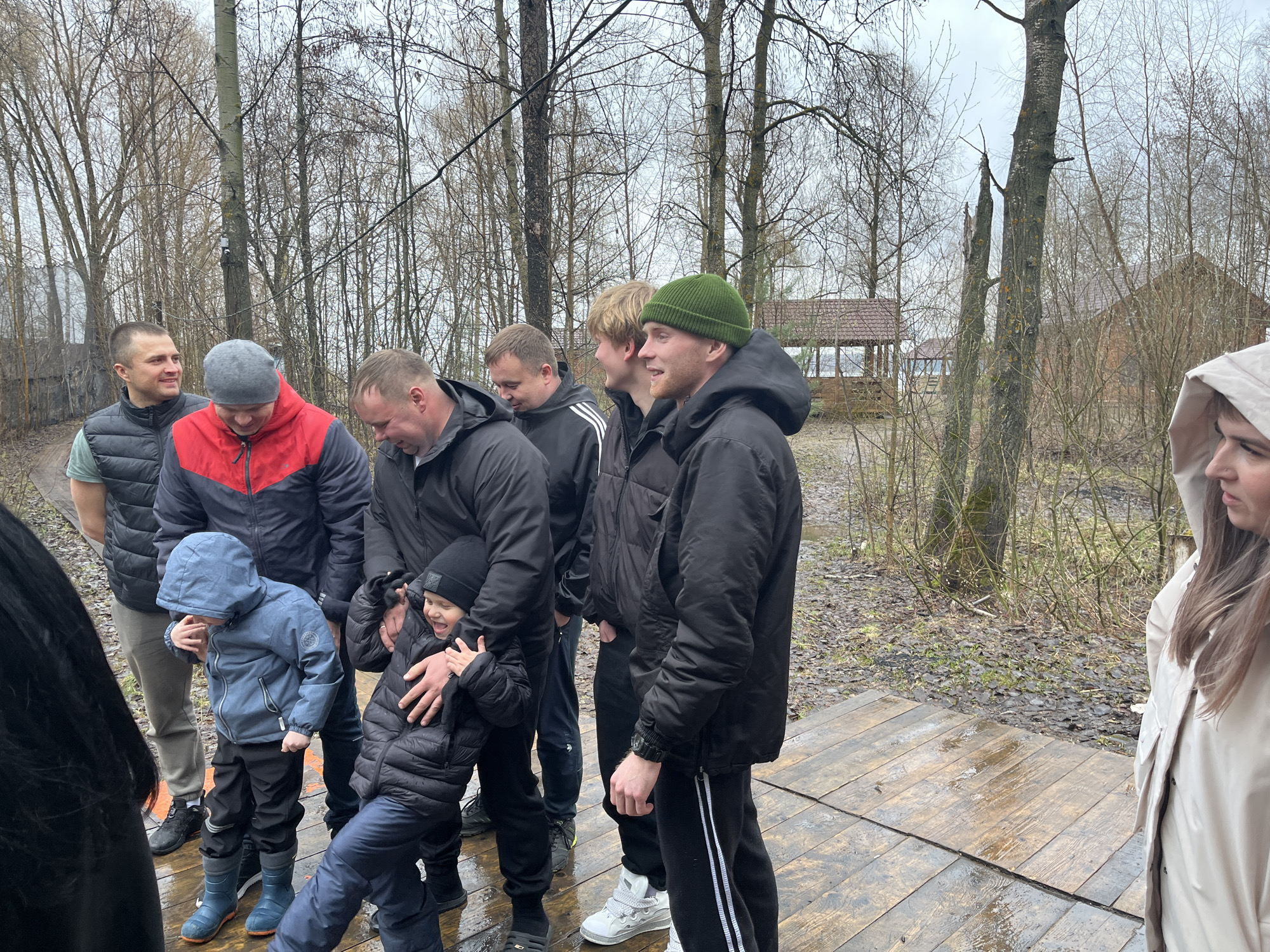 A group of individuals, including children and adults, are gathered on a wooden platform in what appears to be an outdoor setting. The backdrop includes trees and a structure resembling a gazebo. The weather seems overcast and possibly wet, as the platform is damp. Location is near Chaikovsky, Russia.
The people are dressed in various outerwear, suggesting cooler temperatures. Some are wearing hats or hoods. The adults appear to be interacting with the children, potentially engaging in a playful activity. The children are dressed in winter weather gear.
The scene evokes a sense of a family or community gathering in a natural environment.