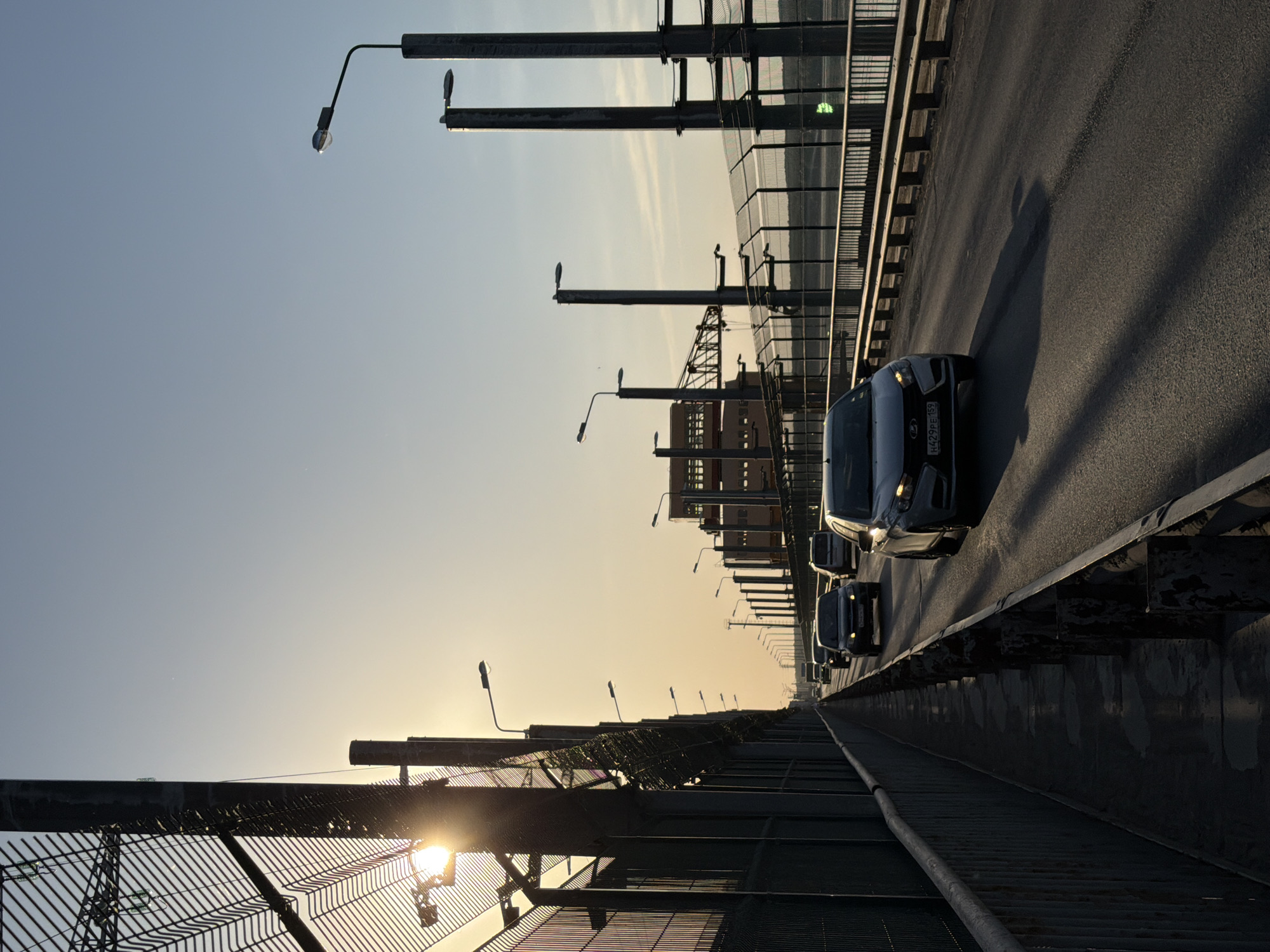 The image shows a view along the Votkinsk Bridge in Chaikovsky, Russia. The view is low, facing along the bridge deck. Multiple black cars are visible in a row, moving forward on the road. There are light posts along the sides of the bridge, with a metal safety fence closer to the foreground and a taller safety barrier on the other side. The sky is light blue and golden yellow at the horizon, suggesting either sunrise or sunset. The lighting conditions suggest that it is daytime.