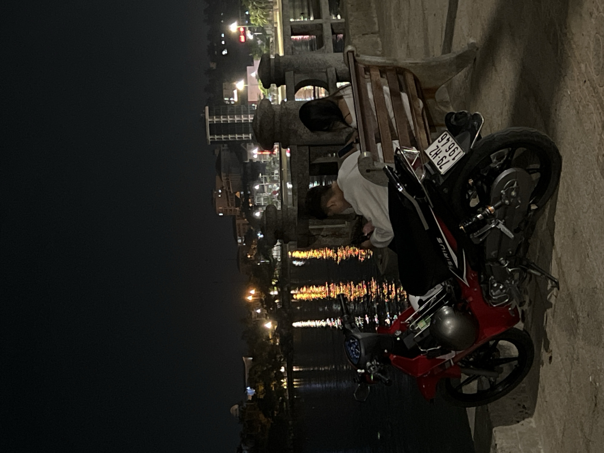 The image depicts a nighttime scene in Bac Nha Trang Ward, Vietnam, beside a body of water. Two individuals are present in the foreground. One person is seated on a wooden bench with robust stone supports, facing away from the camera. Another person is leaning against a red and black motorcycle, looking down at an unseen object, likely a phone.
The motorcycle has "SIRIUS" written on its side and a visible license plate that reads "79-H2 196.46". A dark helmet rests on the seat of the motorcycle. The background features an urban environment with numerous illuminated buildings and streetlights, whose bright reflections are vividly cast upon the dark surface of the water, suggesting a riverside or lakeside location within a city. The overall atmosphere is dark, illuminated solely by artificial lights from the city and streetlamps. The ground appears to be concrete or paved. The scene suggests a casual evening stop or a moment of rest by the waterfront.