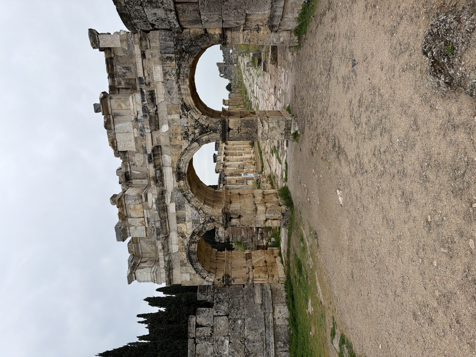 The image shows the ruins of an ancient Roman gateway in Pamukkale, Turkey, with three arches still standing. The structure is made of large, weathered stone blocks, with some vegetation growing on and around it. A dirt path leads through the gateway, with more ruins visible in the background. The sky is overcast.