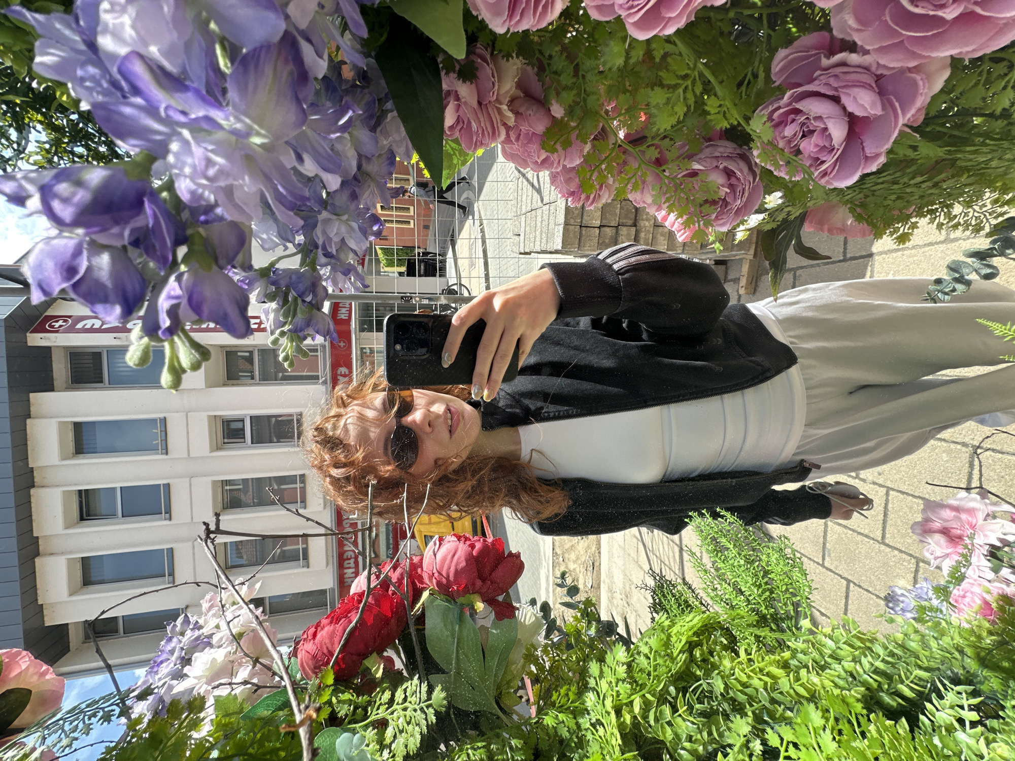 The image captures a selfie of a young woman with reddish-brown curly hair and sunglasses, reflected in a mirrored surface adorned with artificial flowers and greenery. She holds a black smartphone, wearing a black jacket with white stripes on the sleeves, a white top, and light-colored pants. Her fingernails are painted a light, possibly silver, color.
The reflective surface is extensively decorated with artificial flora. Clusters of purple/lavender wisteria-like flowers cascade from above, complemented by pink roses or peonies and some red peonies. Various types of green foliage fill the spaces, creating a lush, framing effect around the woman's reflection.
Behind her reflection, a sunny urban scene is visible, suggesting daytime. Multi-story buildings with numerous rectangular windows line a street, featuring a paved sidewalk. On a red shop sign reflected in the background, Cyrillic text is partially visible, including the letters "ДНК" (DNK) and a white medical cross symbol, indicating a pharmacy or medical-related business. These textual and architectural elements are consistent with an urban setting in Novi Sad, Serbia. The overall mood is bright and casual.