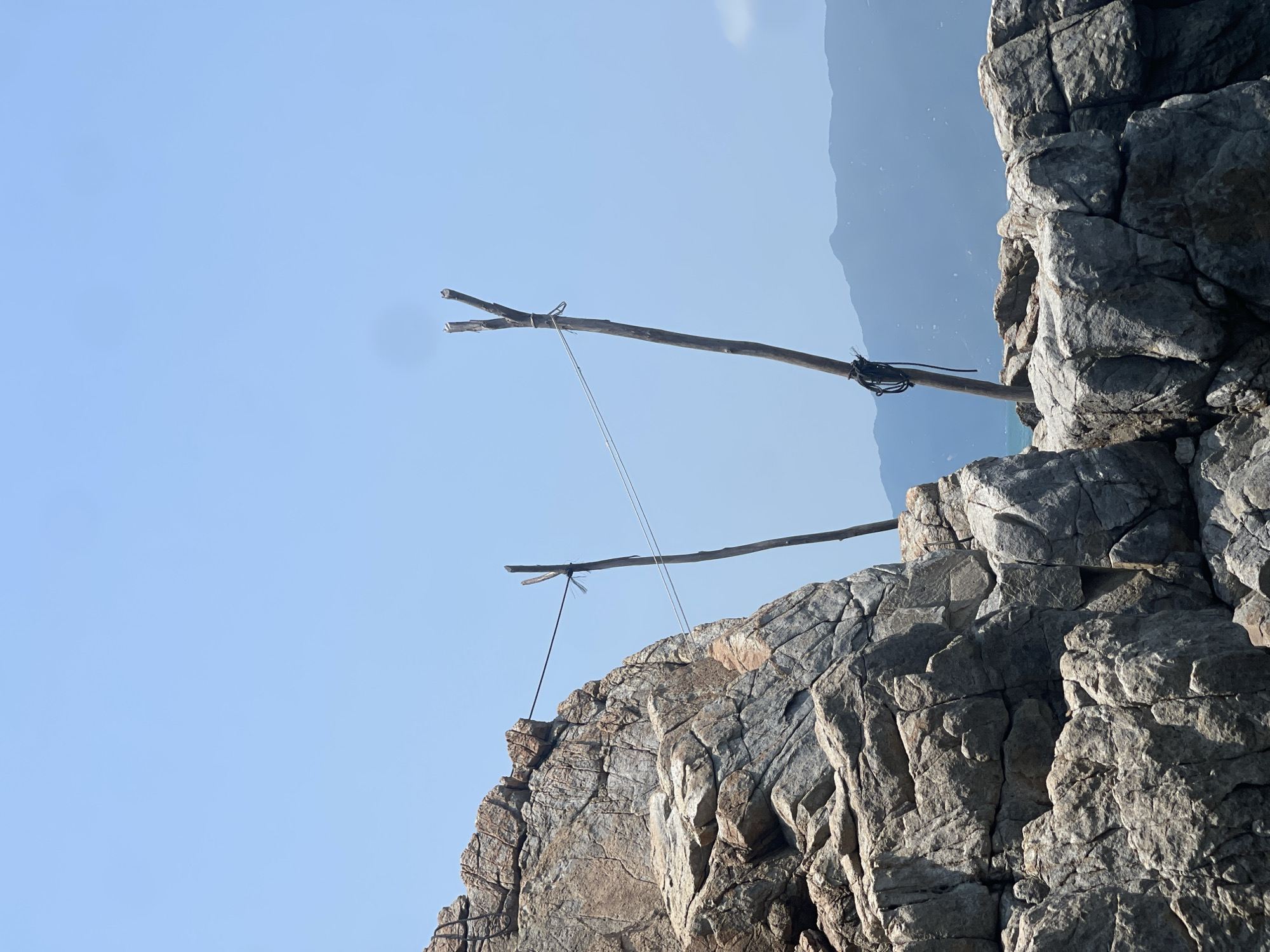 A landscape scene depicts rugged, light gray rocks in the foreground, extending from the bottom to the upper right. Protruding from the rocks are two wooden poles, lashed with black cord and strung with a thin white line. The upper pole is near the upper right, while the lower pole is centered. The background consists of a light blue sky and a hazy mountain ridge in the distance. The image seems to be taken in Vĩnh Lương, Vietnam.