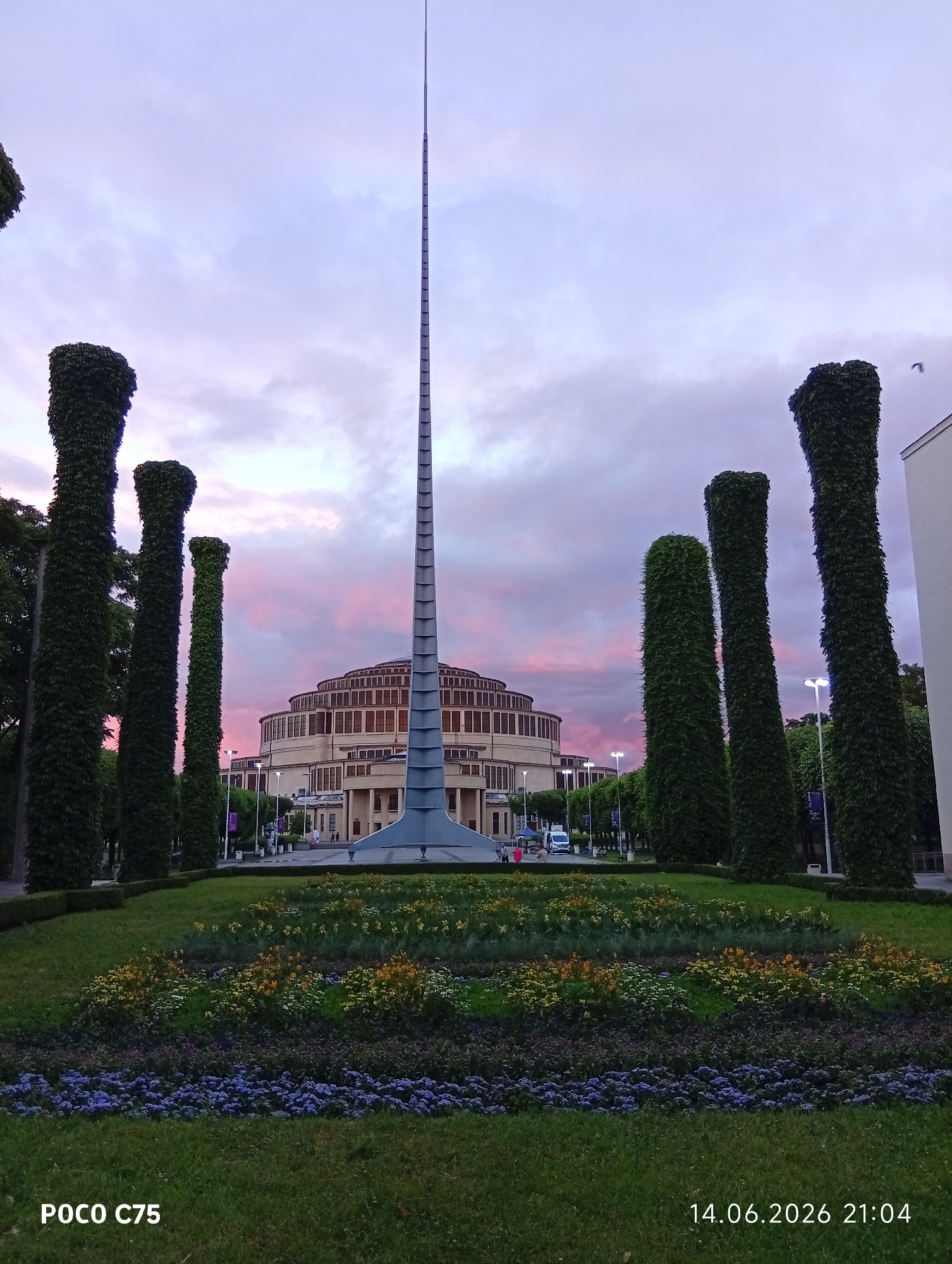 The image captures the Centennial Hall in Wrocław, Poland, at dusk. A tall, slender, metallic spire dominates the foreground, with several large, topiary-like trees on either side. In the mid-ground, the distinctive circular Centennial Hall building is visible, its facade illuminated by the fading light. The sky above is a gradient of soft pink and purple, indicative of sunset. The foreground features a meticulously maintained garden with rows of colorful flowers in bloom, transitioning to lush green grass. The timestamp "14.06.2026 21:04" and "POCO C75" are overlaid on the bottom of the image. A few distant figures and vehicles are present on the pathway in front of the building, suggesting a public space. The overall scene is one of civic grandeur combined with natural beauty.