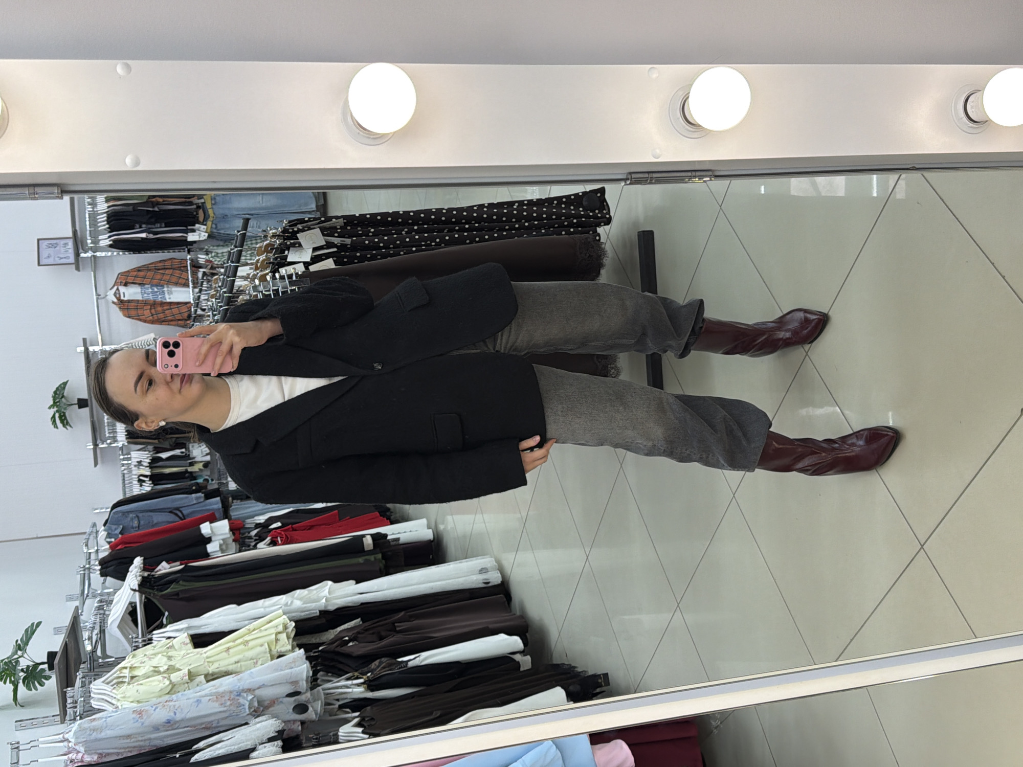 The image appears to be a reflection in a mirror inside a clothing store. A person is taking a selfie, visible by their phone held up in the reflection. The person is wearing a white shirt, a black blazer, grey pants, and dark red boots. Racks of clothing are visible in the background, including dresses, pants, and shirts in various colors and patterns. The floor is tiled with light-colored tiles arranged in a diamond pattern. A row of lights is positioned above the mirror, casting illumination onto the scene. The city of Chaikovsky, Russia, is specified as the location.
