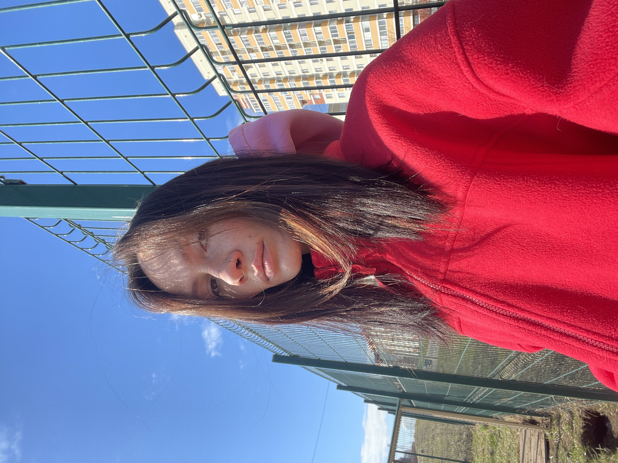 A woman wearing a red hooded sweatshirt stands near a green metal fence, looking at the camera. The background features a blue sky with a few clouds and a tall apartment building in the distance. The location is likely an urban area in Izhevsk, Russia, given the apartment building and the presence of a metal fence typically found in city parks or public spaces. The woman's hair is dark, and she appears to have a nose ring. The photo appears to be taken in daylight, with a clear view of the sky and surrounding elements.