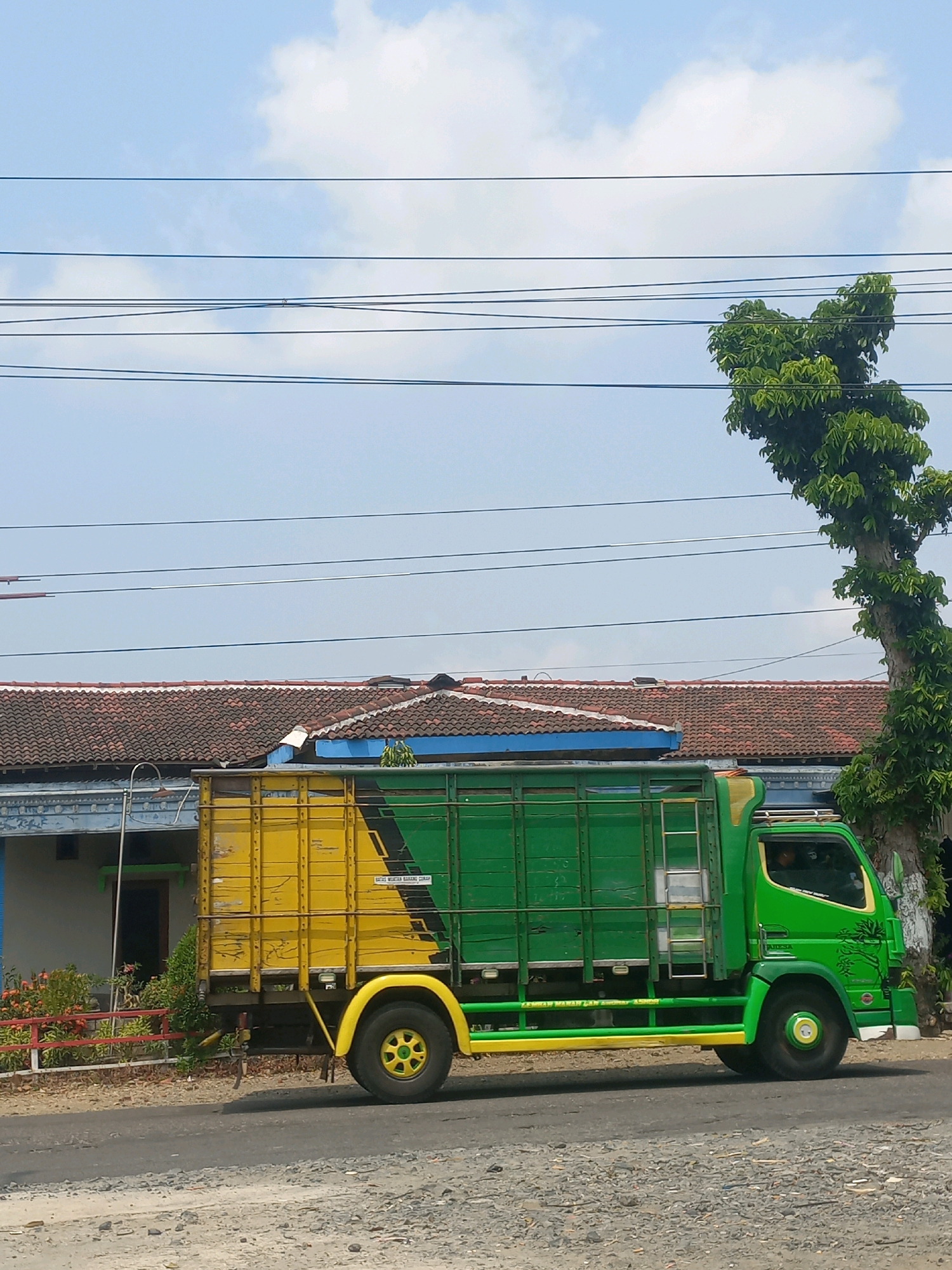A bright green and yellow truck is parked on an asphalt road in front of a building with a tiled roof. The truck has a tall, enclosed cargo area that is predominantly green with a yellow section on the left side. A ladder is attached to the side of the cargo area. The truck's cab is green, and a person is visible inside. A large tree with lush green leaves stands to the right of the truck. Power lines are strung across the top of the image. The sky is blue with scattered clouds. The scene appears to be in a rural or semi-urban area of Baturetno, Indonesia.