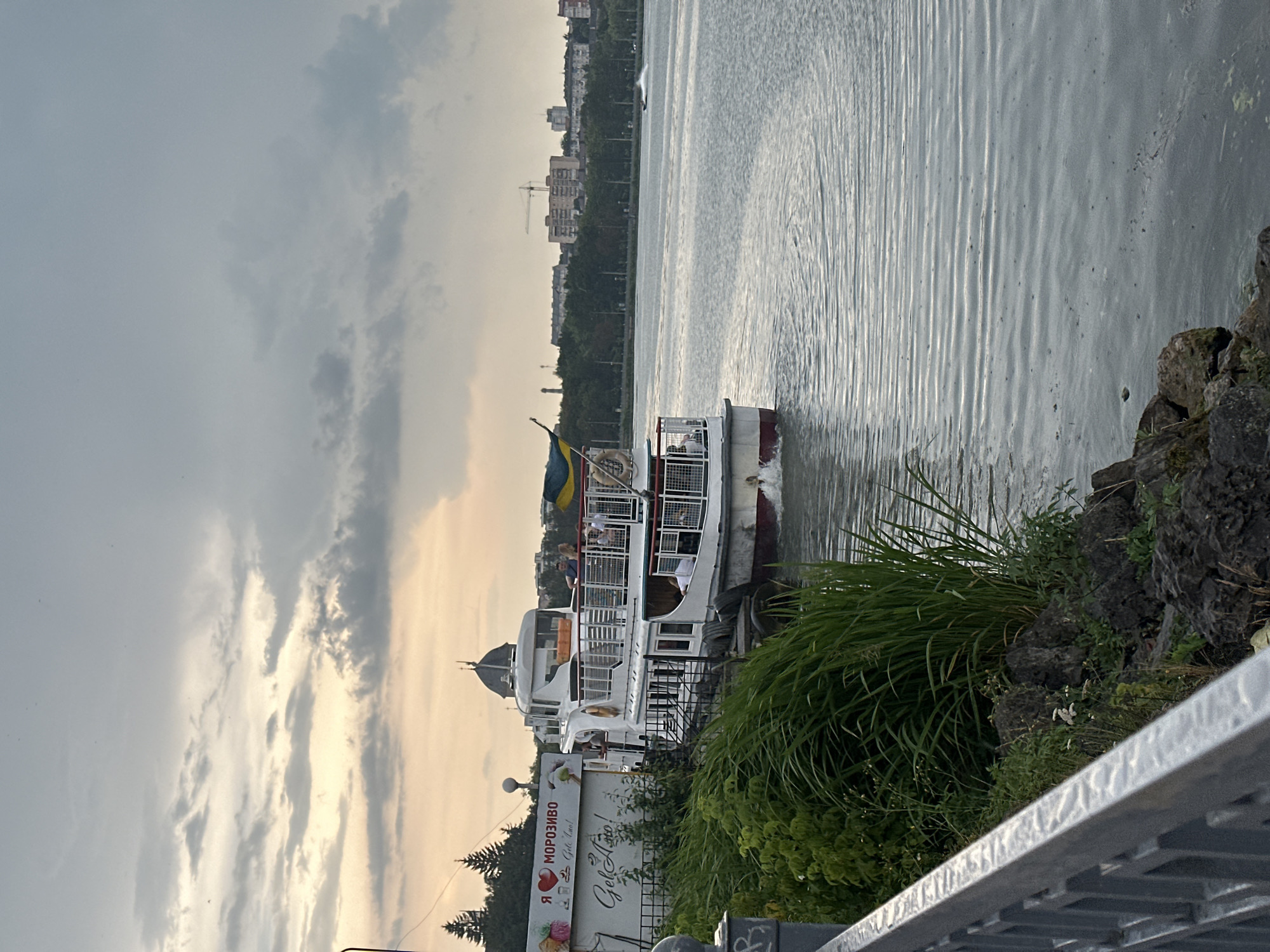An overcast sky looms over a wide body of water, possibly a lake or river, in Ternopil, Ukraine. In the foreground, a metal railing runs along the edge of the embankment, which is covered with lush green grass and dark rocks. A white passenger boat, with a Ukrainian flag flying from its stern, is docked or moving slowly near the embankment. People are visible on the upper deck of the boat. On the shore, a white sign with red lettering and images of ice cream, as well as text in Ukrainian that translates to "I love ice cream" and "Gelato!", is visible. In the distance, a tree-lined embankment is dotted with buildings, including what appears to be a church dome. A construction crane is also visible in the far distance. The water is rippled, indicating gentle movement. The overall scene suggests a late afternoon or early evening setting.
