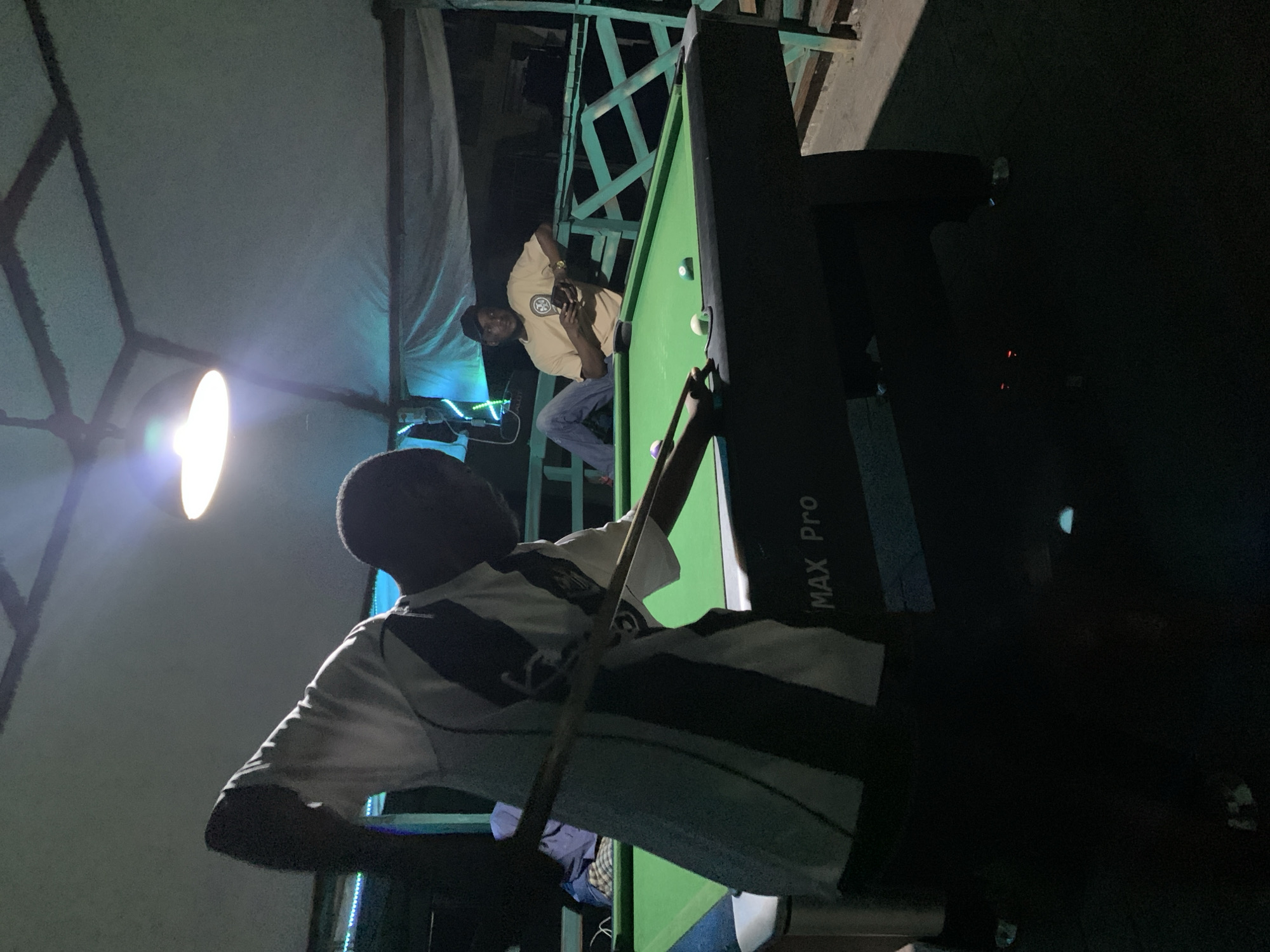 The image depicts two individuals at a pool table in an outdoor, shaded area, possibly a bar or recreational spot, located in Lokoja, Nigeria. The primary subject, in the foreground and slightly to the left, is a man in silhouette, wearing a black and white striped shirt, in the process of playing pool. He is bent over the table, holding a cue stick with both hands, aiming at a white cue ball. His posture suggests an active play. To the right and further back, a second man is seated at the edge of the pool table, wearing a tan t-shirt and blue jeans, looking down at his phone and holding it in his right hand. The pool table is green with several balls visible, including a white cue ball, and other colored balls scattered on its surface. The environment is dimly lit, with an overhead light fixture providing illumination above the table. The background shows a rustic, possibly wooden, structure with a light-colored tarp or roof overhead, and a railing. The overall ambiance is casual and recreational.