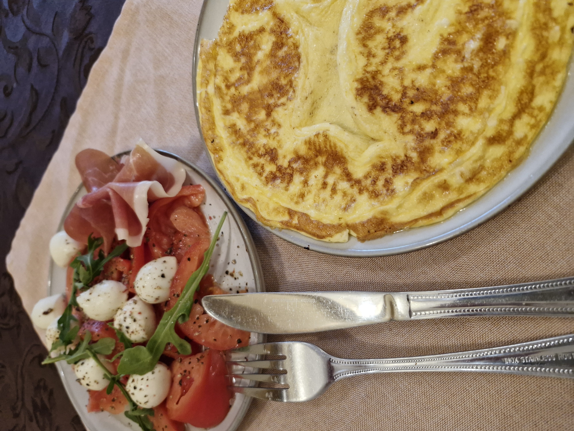 Two plates of food are presented on a light brown fabric tablecloth. A darker, intricately patterned fabric, possibly a placemat or secondary tablecloth, is visible in the upper background.
On the upper left, a round, light-colored ceramic plate holds a caprese-style salad. This includes sliced red tomatoes, numerous small white spherical mozzarella balls heavily speckled with black pepper, and scattered fresh green arugula leaves. To the right side of this plate, several thin slices of folded prosciutto, displaying a light pink color with subtle fat marbling, are arranged.
In the lower right, a second round, light-colored ceramic plate contains a large, golden-yellow omelette. The omelette exhibits significant browning and a folded texture, indicating thorough cooking.
Positioned between the two plates in the foreground are a silver-colored metal fork and knife, lying parallel on the tablecloth. Both utensils feature a decorative beaded pattern along the edges of their handles. The fork has four tines, and the knife has a straight blade.
The scene is illuminated by an overhead or frontal light source. The specific indoor location, city, and country are unknown.