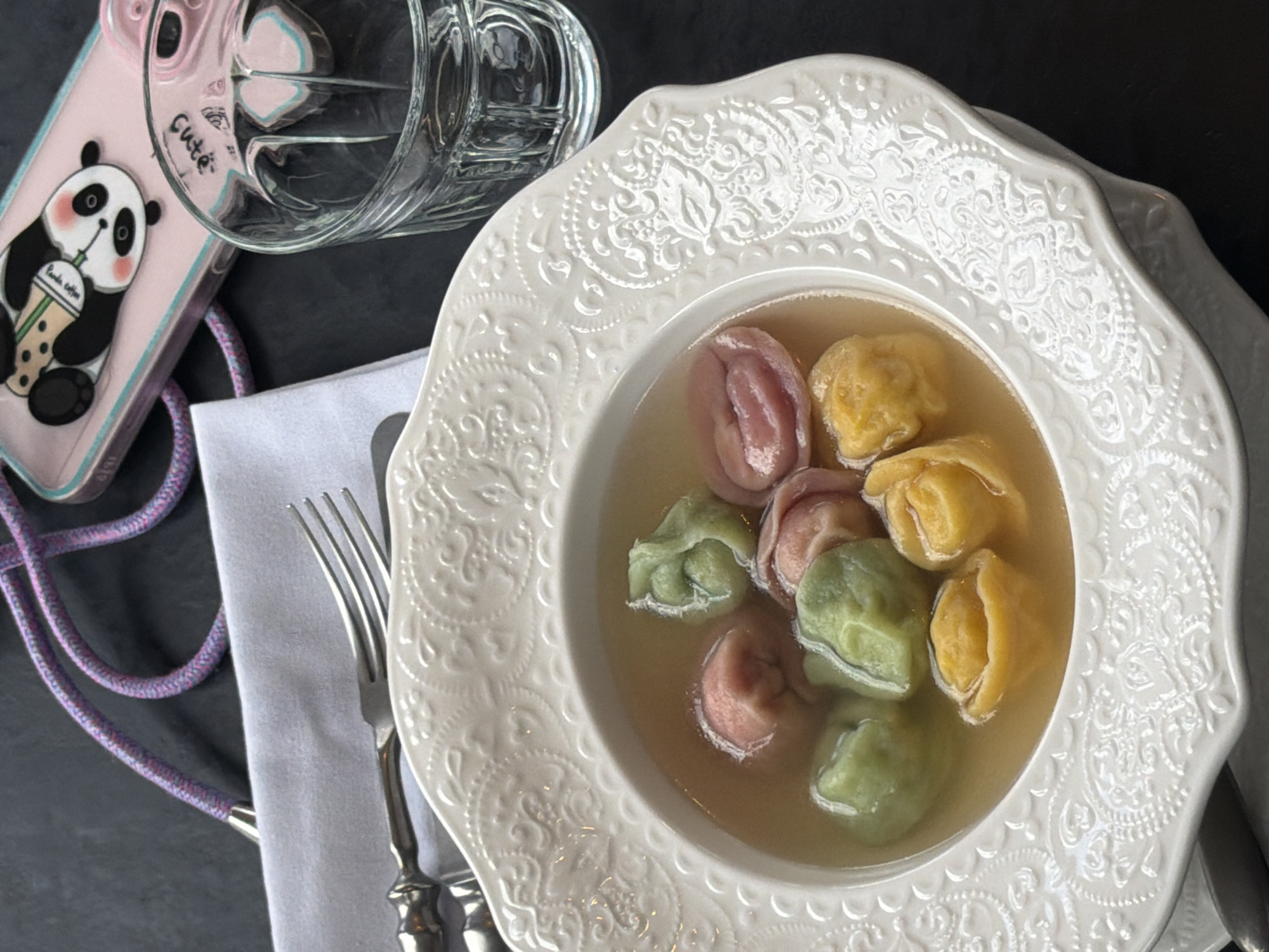 A high-angle, close-up shot features a white, ornate bowl filled with colorful broth and dumplings on a dark surface. The dumplings are green, yellow, and pink. A glass of water rests above the bowl next to a pink phone case with a panda graphic. Silverware is positioned on a folded white napkin. The scene evokes a dining setting, and the food suggests a cultural dish. The lack of specific architectural cues, distinct landmarks, or additional location-specific elements prevents a confident conclusion about Krasnoyarsk, Russia.