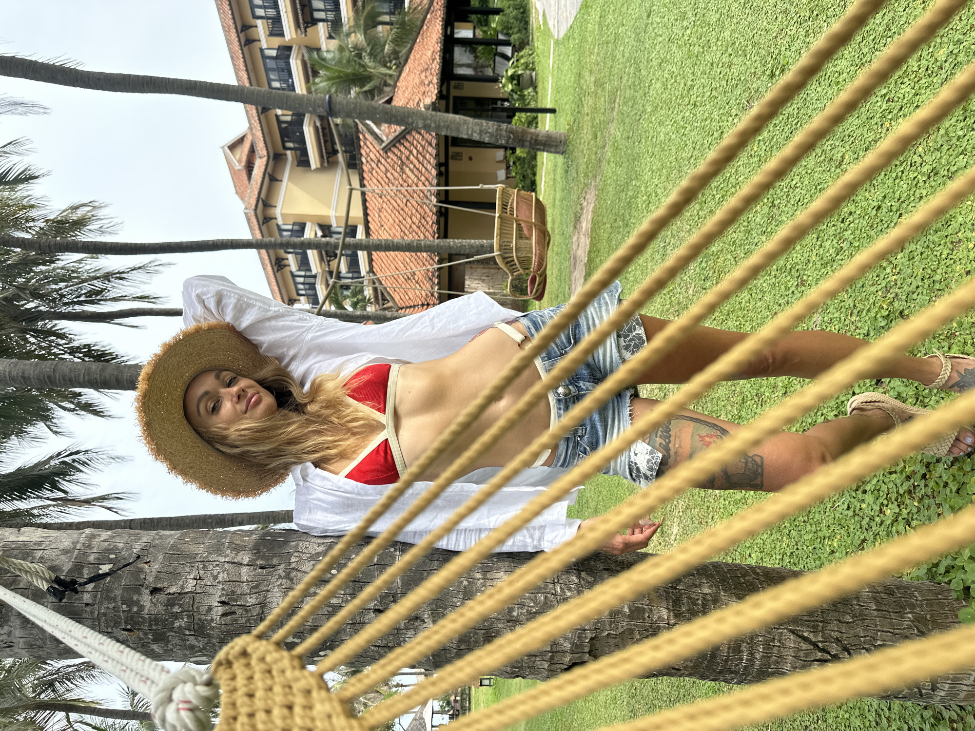 A woman lies in a hammock tied between two palm trees in Phu Thuy Ward, Vietnam. She wears a red bikini top, a white unbuttoned shirt, jean shorts, and a straw hat. The hammock is made of thick, light brown rope. The background features a green lawn, more palm trees, and a building with a red-tiled roof and yellow walls.