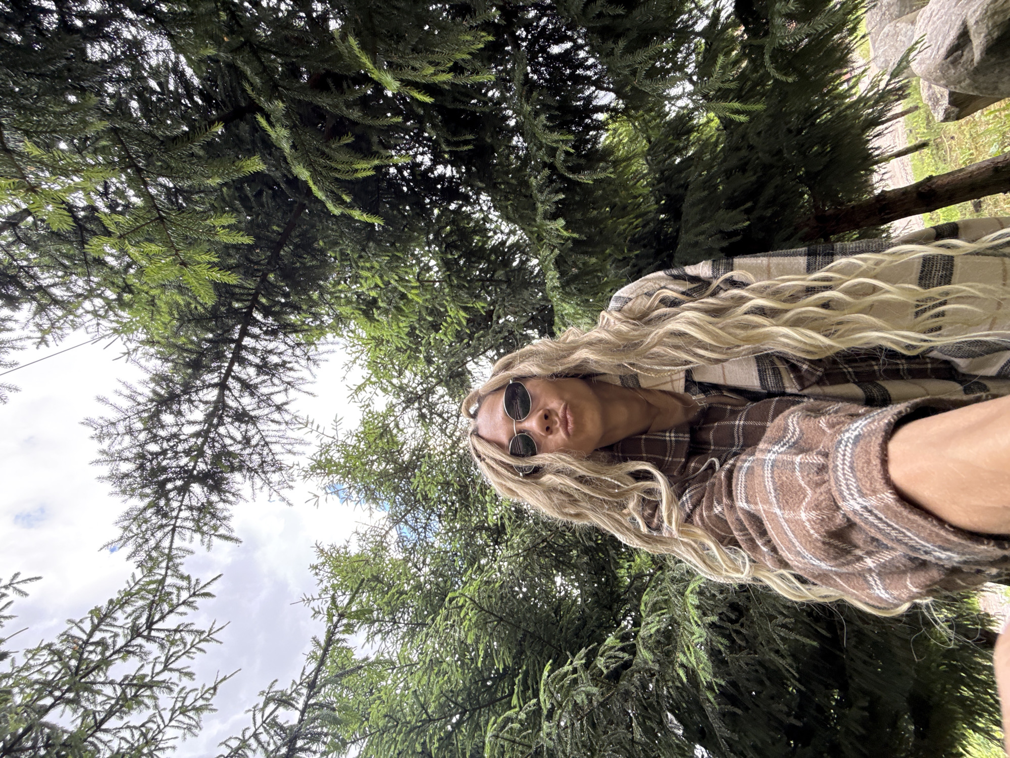 A low-angle shot shows a person with long, blonde dreadlocks and round sunglasses, wearing a brown plaid shirt, against a backdrop of dense green fir trees. The sky is visible through the branches. The location is outdoors, seemingly in a wooded area. The specific location within Rusanovo, Russia, cannot be determined from the image.