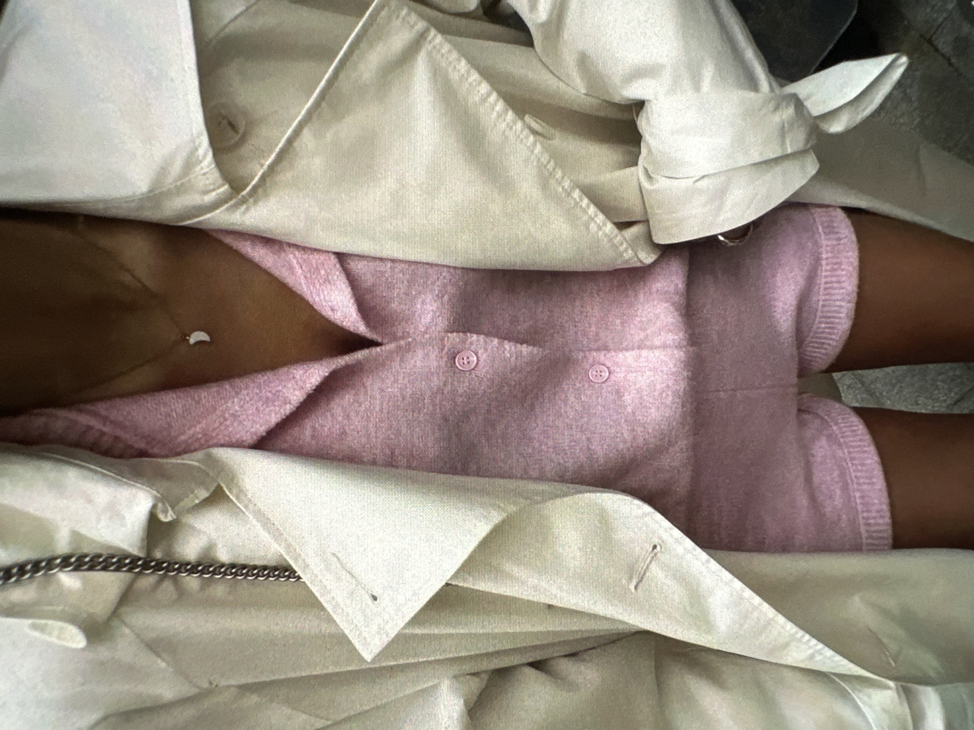A close-up overhead shot presents a person wearing a pink short-sleeved top with two buttons, partially obscured by a beige trench coat. A silver chain is visible on the lower left edge of the coat. The person also appears to be wearing a necklace. No location in Yerevan, Armenia can be determined from the image.
