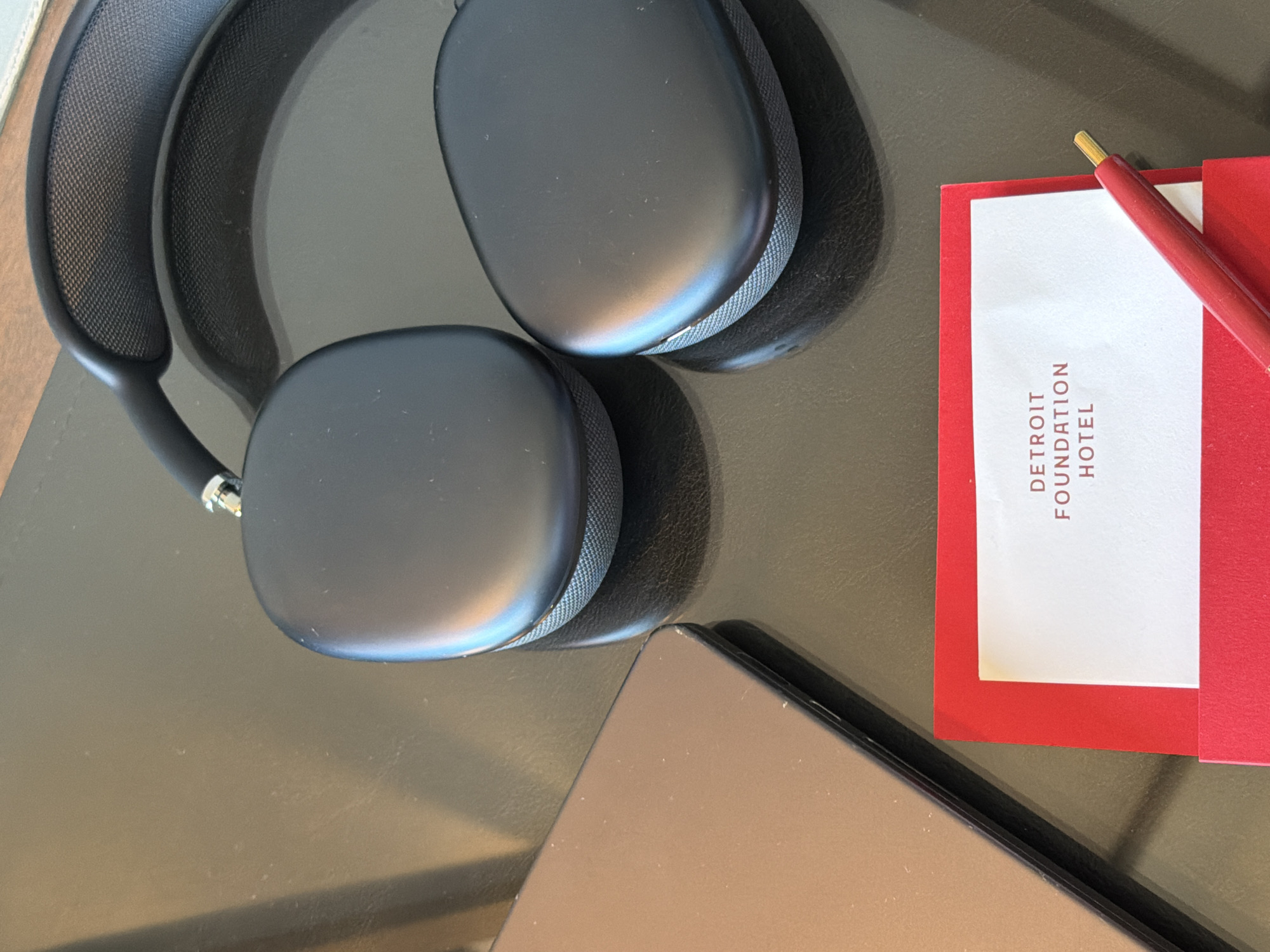 On a dark, reflective surface, possibly a table, are a pair of black over-ear headphones, a closed metallic laptop, a red envelope with a white card inside, and a red pen. The card within the envelope is printed with the text "DETROIT FOUNDATION HOTEL" in red capital letters. The headphones appear to be the Apple AirPods Max. The laptop is dark brown or bronze. The scene is lit from above, casting shadows on the surface. The background is blurred.