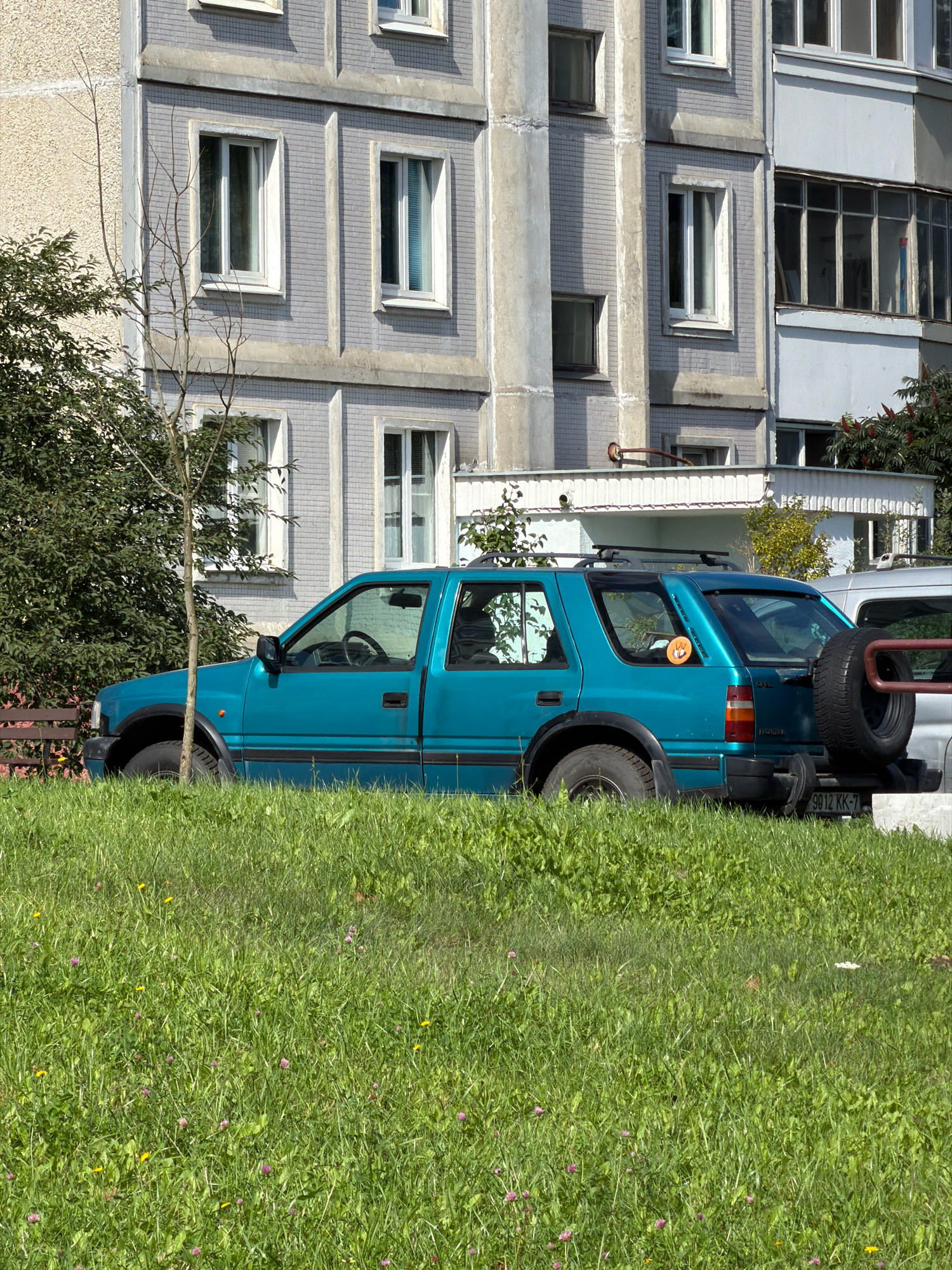 A turquoise SUV is parked on a grassy area in front of a grey, multi-story apartment building. The SUV has a spare tire mounted on the rear and a sticker of an orange creature with horns on the back window. A portion of a silver vehicle is visible to the right of the SUV. The building has numerous windows, some with white frames and blinds. The grassy area in front of the vehicles is somewhat overgrown and dotted with small pink and yellow flowers. The scene appears to be in an urban residential area.