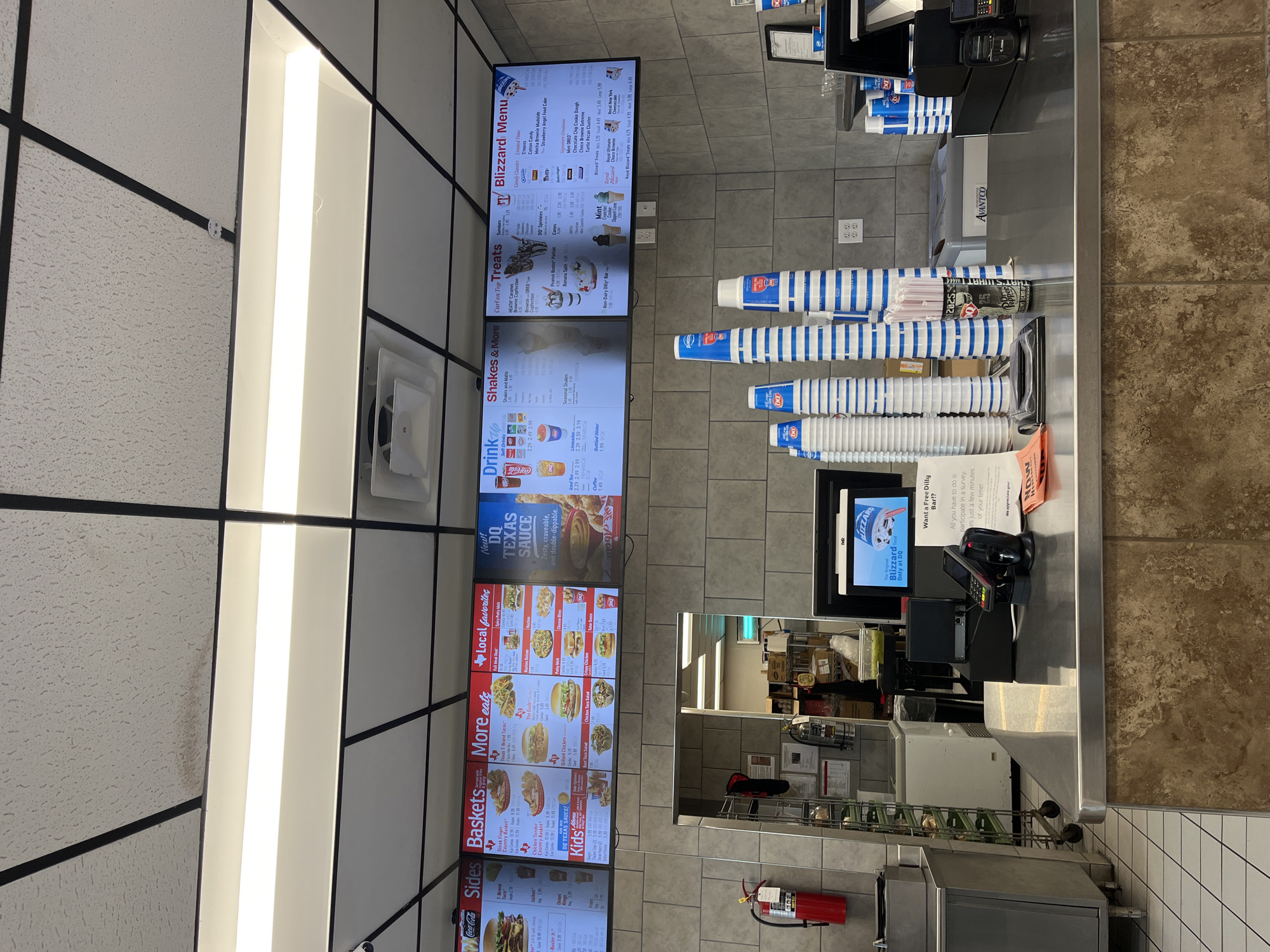 This is an interior shot of a Dairy Queen restaurant in Johnson City, Tennessee, United States of America. The image focuses on the ordering counter area. Above the counter are digital menu boards displaying "Blizzard Menu", "Treats", "Shakes and Malts", "Drinks", "More Eats", "Baskets", and "Sides". The menus feature prices for various Dairy Queen items. Several stacks of Dairy Queen branded cups are on the counter. A cash register, a customer-facing screen displaying the "Blizzard" logo, and a payment terminal sit on the counter. A poster indicating a "Free Dilly Bar" incentive is visible. Behind the counter is a partial view of the kitchen area, including shelving with supplies and a fire extinguisher. The general color palette is neutral with tile walls and a textured ceiling with recessed lighting.