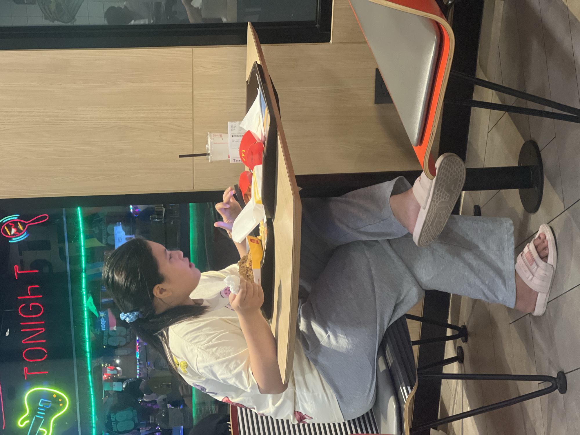 A woman sits at a table in a McDonald's restaurant. She is wearing a light-colored t-shirt with a pattern of flowers and grey sweatpants. She has her hair pulled back in a ponytail and is holding a piece of food in her right hand while looking at her phone. A McDonald's meal is on the table in front of her, including a tray with a drink cup, French fries, and a burger. The restaurant has a neon "TONIGHT" sign visible in the background. The table is made of wood, and the chairs are orange and grey. The floor is tiled.
