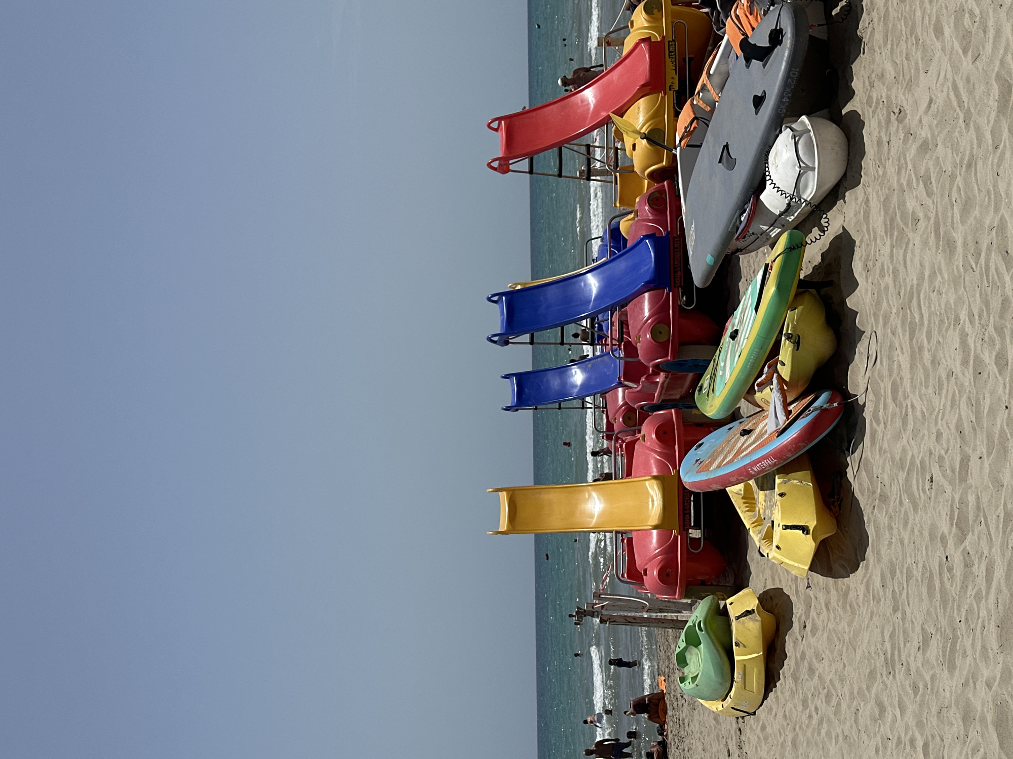 The image depicts a beach scene with several recreational watercraft and pedal boats lined up on the sand. In the background, the ocean is visible with several people swimming and enjoying the water. The sky is clear and blue.
Specifically, the foreground shows various types of boats:
- Several brightly colored pedal boats, shaped like cars, are positioned in the middle ground. Some of these have slides attached.
- Two red pedal boats are in the front row.
- Yellow pedal boats are also present, some with green kayaks or paddleboards resting on them.
- A gray surfboard and a white flotation device are on the sand in the front right.
- A green kayak is on top of a yellow boat in the front left.
- A paddleboard with red and blue accents is leaning against a yellow boat.
In the middle ground and background:
- The ocean is a vibrant turquoise color with gentle waves.
- People are scattered throughout the water, swimming and wading.
- The beach is made of light brown sand.
- The horizon line is clear, separating the sea from the sky.
The overall atmosphere is that of a sunny day at a beach resort, with water activities readily available. The objects are arranged in a somewhat cluttered but organized manner, suggesting they are rentals or part of a beach service.
