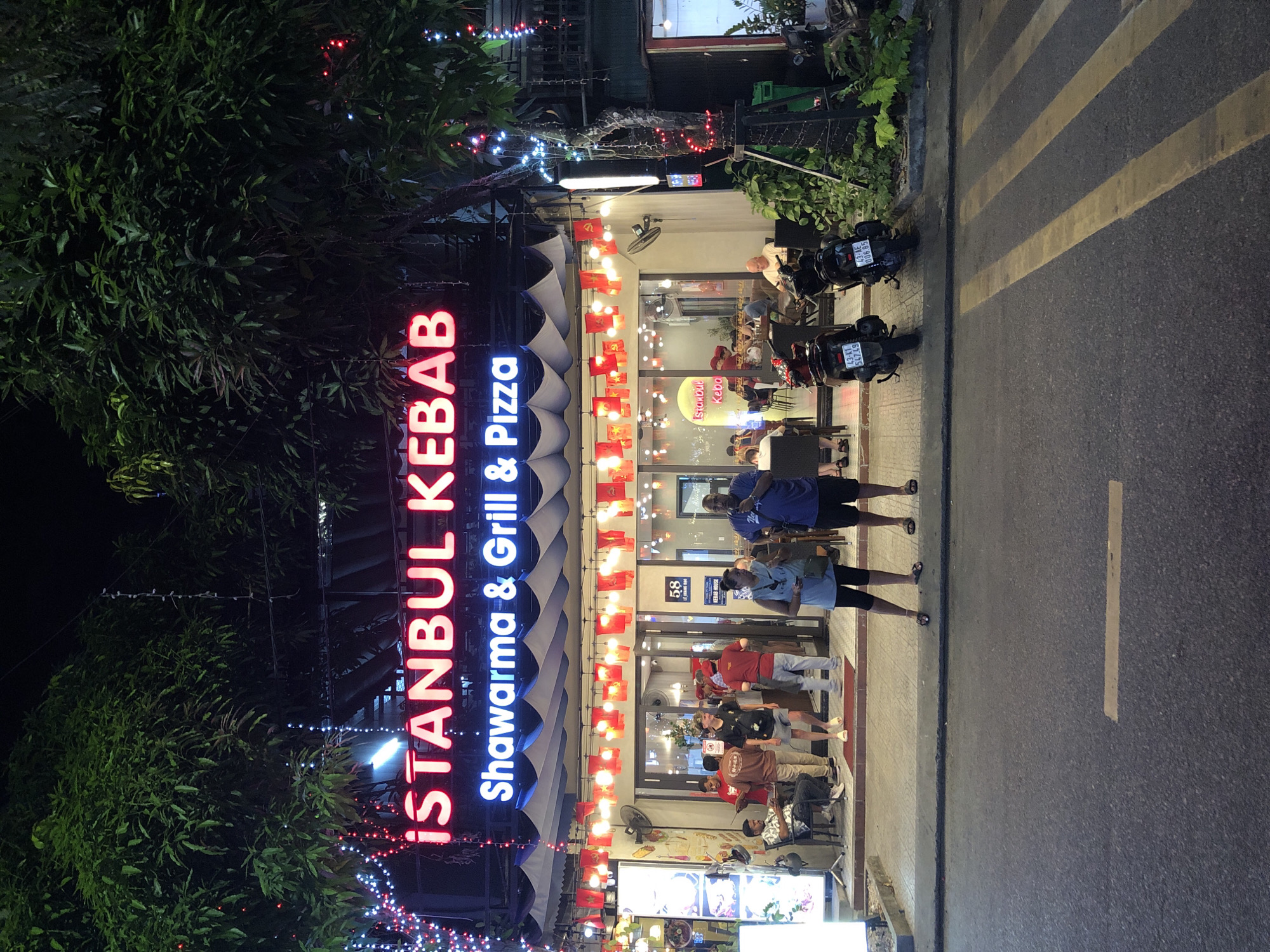 The image shows the exterior of an "ISTANBUL KEBAB Shawarma & Grill & Pizza" restaurant at night, located at 58 Lê Duẩn Street in Đà Nẵng, Vietnam. The restaurant has a neon sign with the name of the establishment. Red and white string lights are draped over trees and the building's facade. Several people are visible standing outside the restaurant and some are seated inside. Two motorcycles are parked in front of the restaurant on the sidewalk. The street in front of the restaurant is paved with asphalt and has yellow markings for a crosswalk. The overall scene depicts a typical urban streetscape at night with a focus on a food establishment.