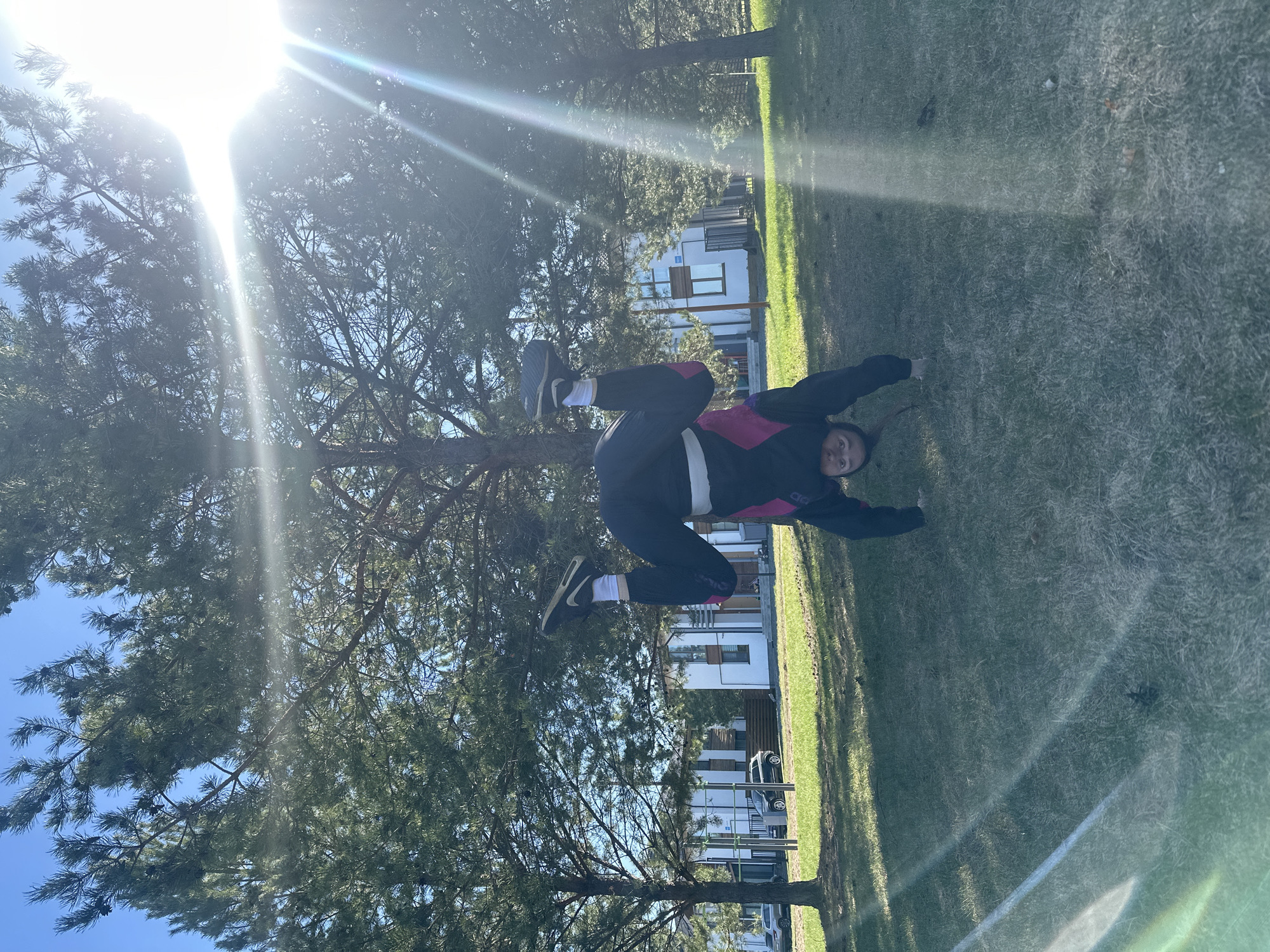 A person is performing a handstand outdoors on a grassy lawn. The person is wearing dark pants, a pink and black jacket, white socks, and dark shoes. In the background, there is a pine tree, a bright sky with sun glare, and a building. The location is Русский Вожой, Russia.