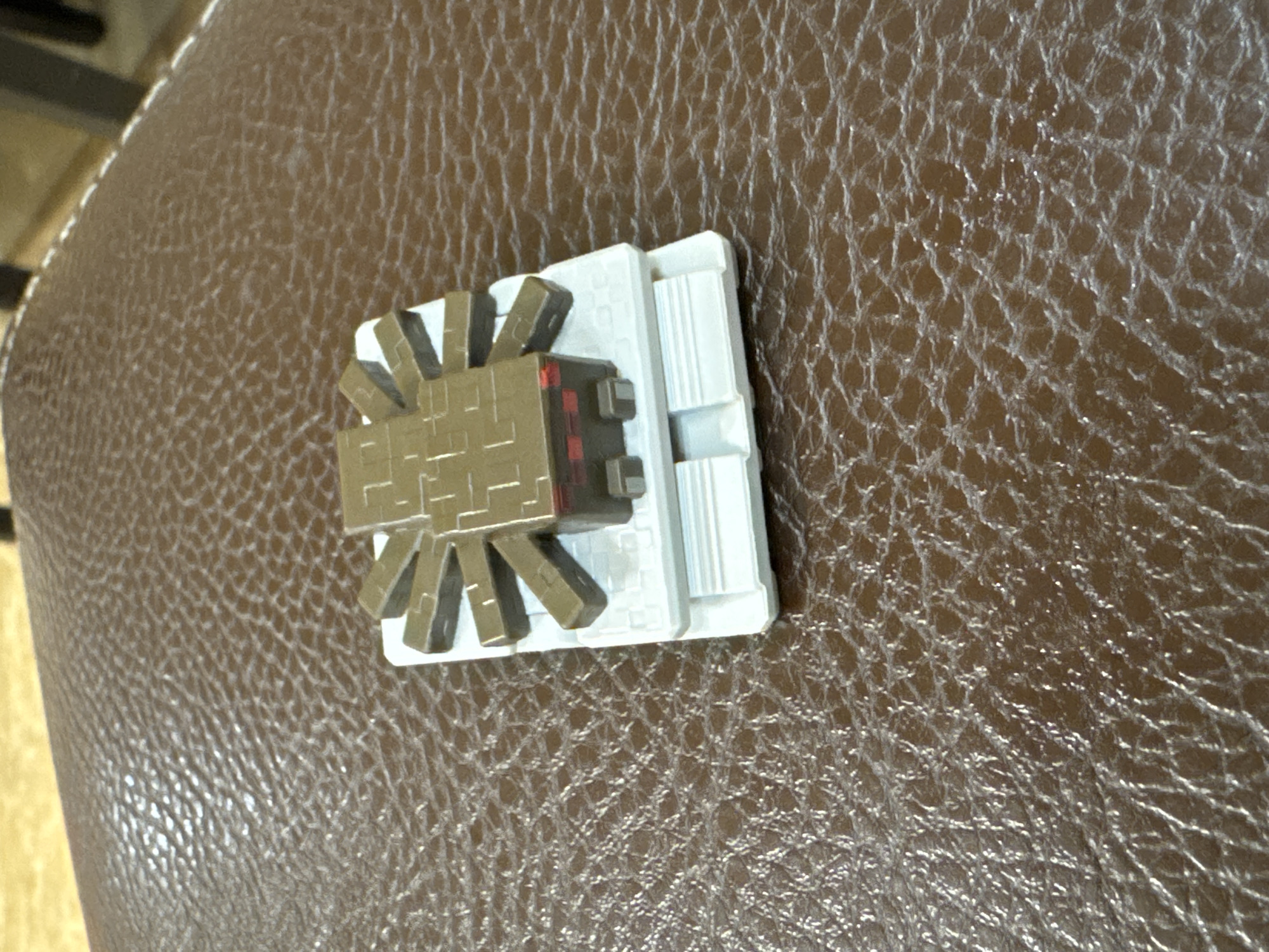 A Minecraft spider toy sits on a brown, textured surface resembling leather. The toy is composed of a brown, blocky spider body with a red eye, attached to a white platform. The platform has ridges on one side. The setting appears to be indoors. Yerevan, Armenia is not evident in this image.