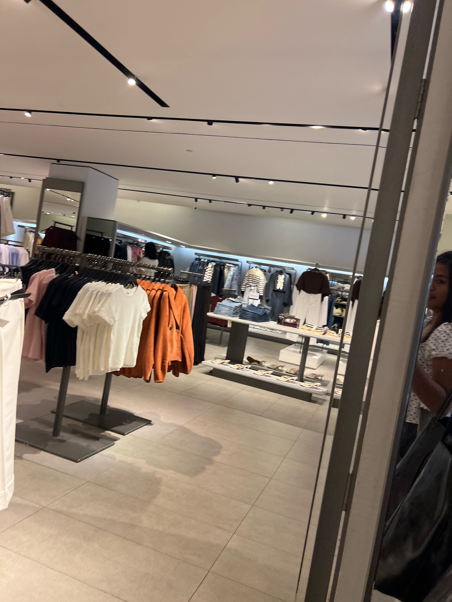 The image shows the interior of a clothing store, likely a Zara, in Kuta, Indonesia. In the foreground, a clothing rack displays several t-shirts and an orange jacket. To the right, a partial reflection of a young woman in a white and black polka dot shirt is visible in a large mirror. The store's tiled floor and white ceiling with track lighting are also visible. In the background, more clothing racks and display tables filled with garments are present, with a glimpse of another shopper or employee further in the store. The overall impression is of a well-lit, modern retail environment.