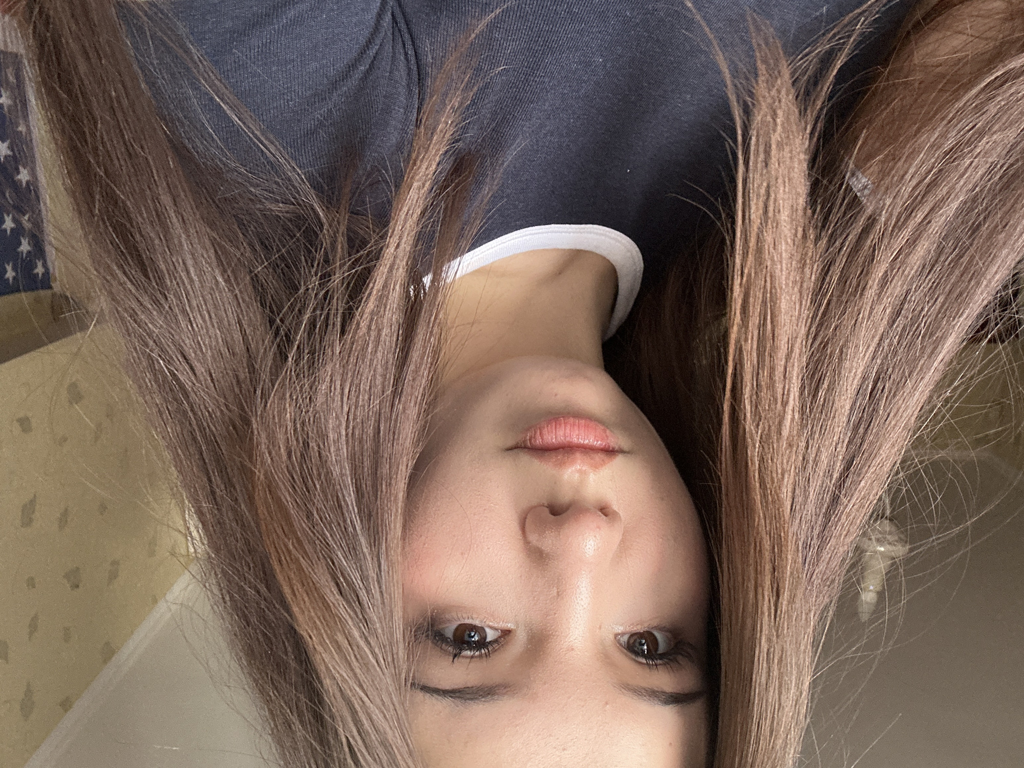 The image shows a close-up, selfie-style portrait of a young woman with long, brown hair. She is wearing a dark t-shirt with a white collar. Her expression is neutral. The background appears to be a room with textured wallpaper and a light fixture partially visible on the left. On the right, a portion of an American flag is visible. There are no discernible landmarks or specific details in the image that would indicate a location in Naberezhnye Chelny, Russia.