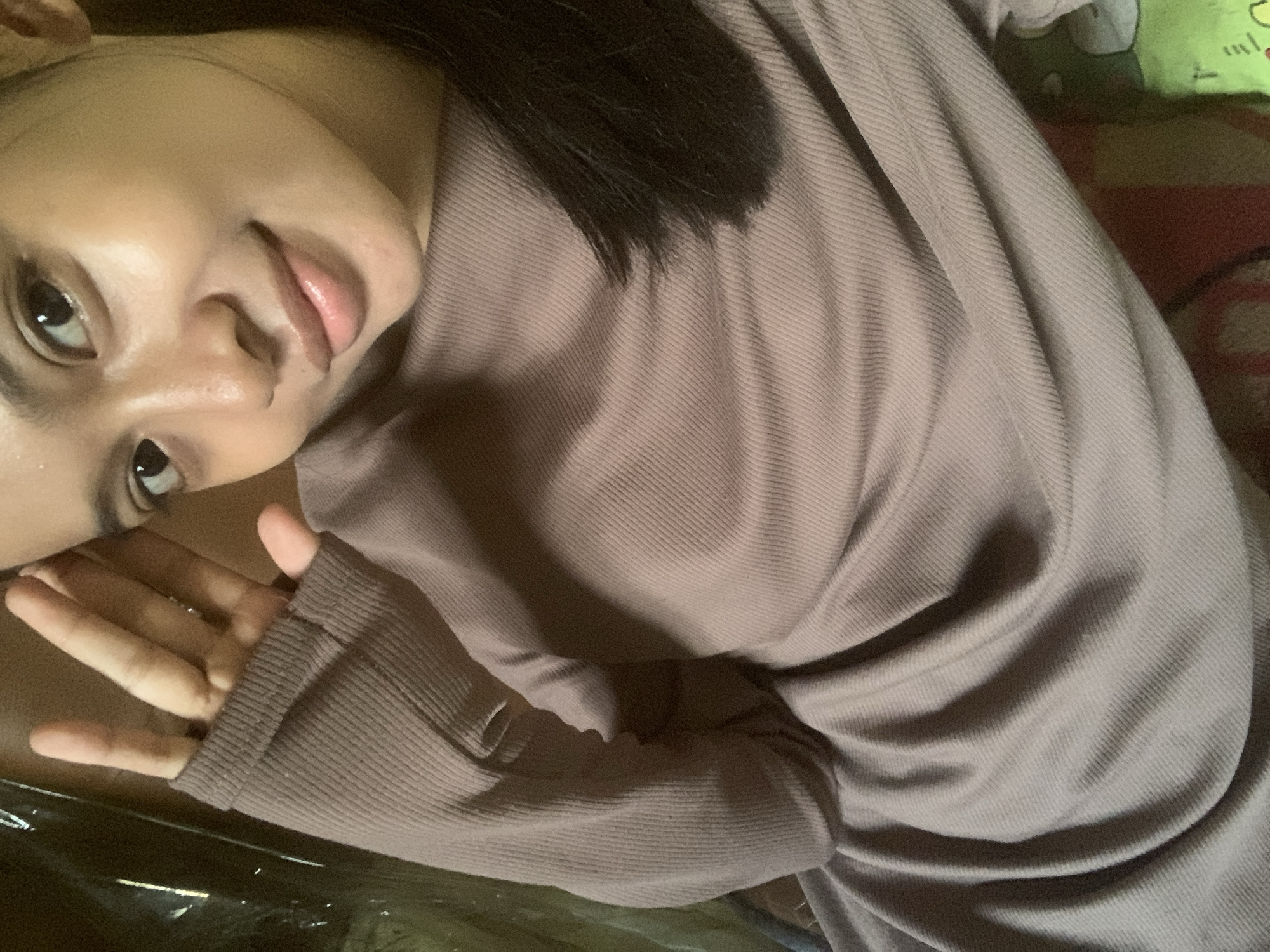 A close-up, high-angle selfie of a young woman is shown. She is wearing a mauve, ribbed long-sleeved shirt. Her right hand is raised to her forehead, obscuring part of her face. Her dark hair falls over her shoulder. The background is out of focus, with a hint of a patterned blanket visible at the bottom right. The lighting suggests an indoor setting. The image does not provide any information that could be used to determine its location in Sungai Meriam, Indonesia.