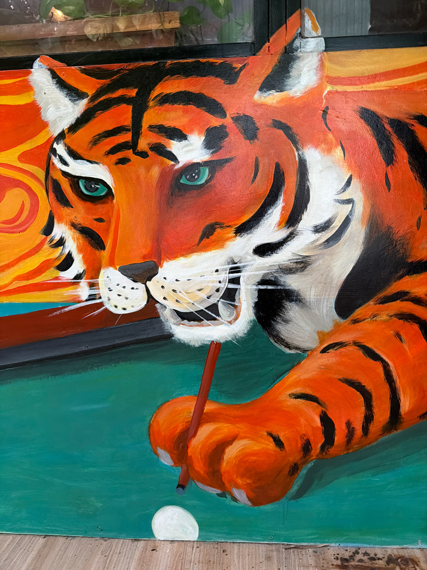 A painting of a tiger's head and paw is depicted. The tiger has green eyes and is positioned as if playing pool. It is holding a pool cue with its paw and aiming for a white ball on a green surface. The background features abstract orange and yellow swirls. The painting is framed by dark wood. There are no people or animals present other than the painted tiger. This artwork is located in Phú Quốc, Vietnam.