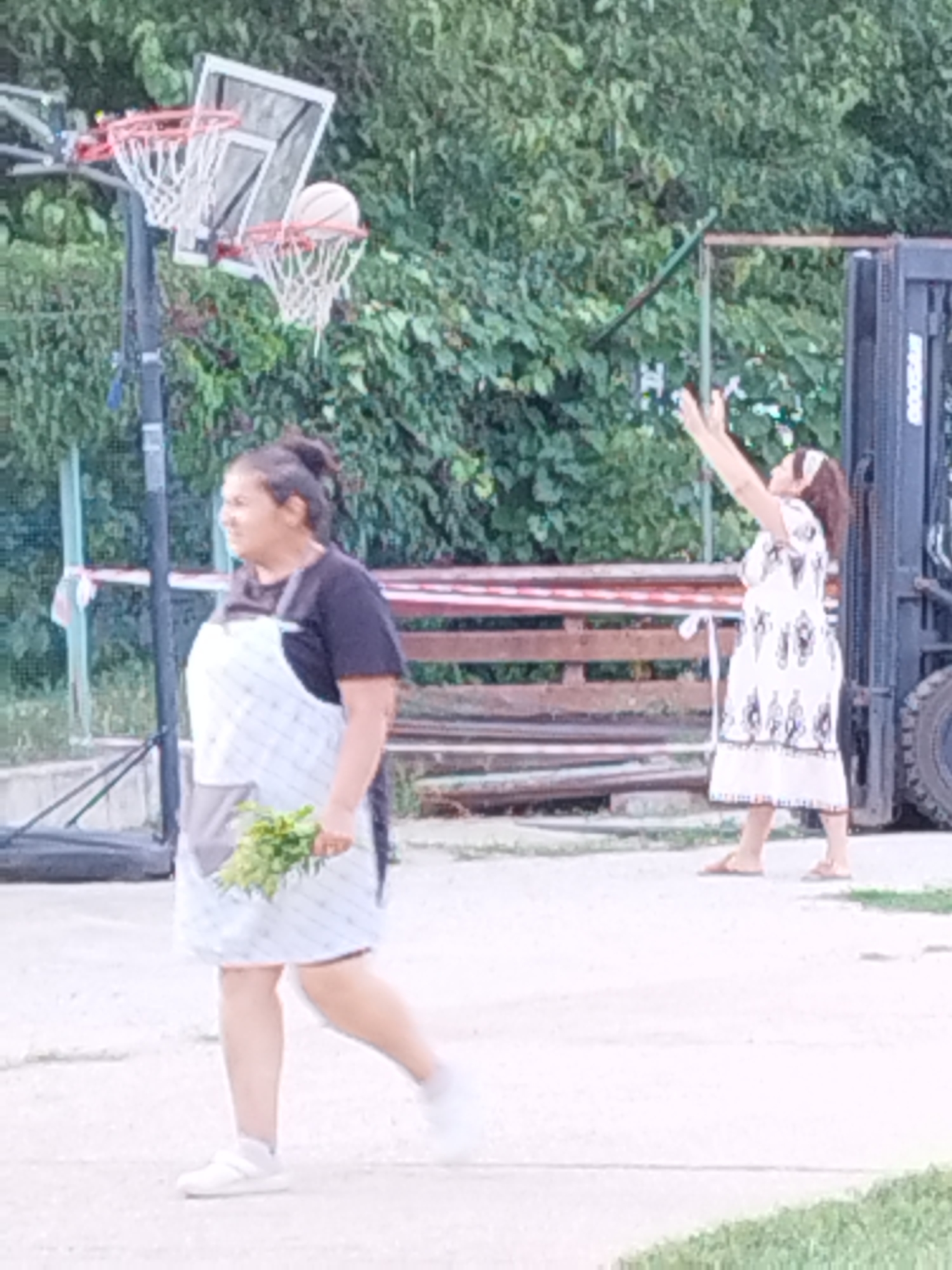 An outdoor scene, likely a casual gathering or event, features two women. In the foreground, a woman wearing a dark t-shirt and a checkered apron is holding a bunch of green leafy herbs and walking towards the left. She has dark hair pulled back and is wearing white sneakers. In the background, to the right, a woman in a white dress with black embellishments and a headscarf is facing away from the camera with her arms raised, possibly in mid-action of throwing something or in a celebratory gesture. Behind her, a large piece of industrial equipment, resembling a forklift, is partially visible. A basketball hoop with a ball at the rim is set up behind the foreground woman, suggesting a possible activity or the presence of a sports area. Wooden structures and dense green foliage form the backdrop. The ground appears to be a paved surface. The scene is not clearly identifiable as being in Bucharest, Romania, but the overall atmosphere suggests a community or recreational space.