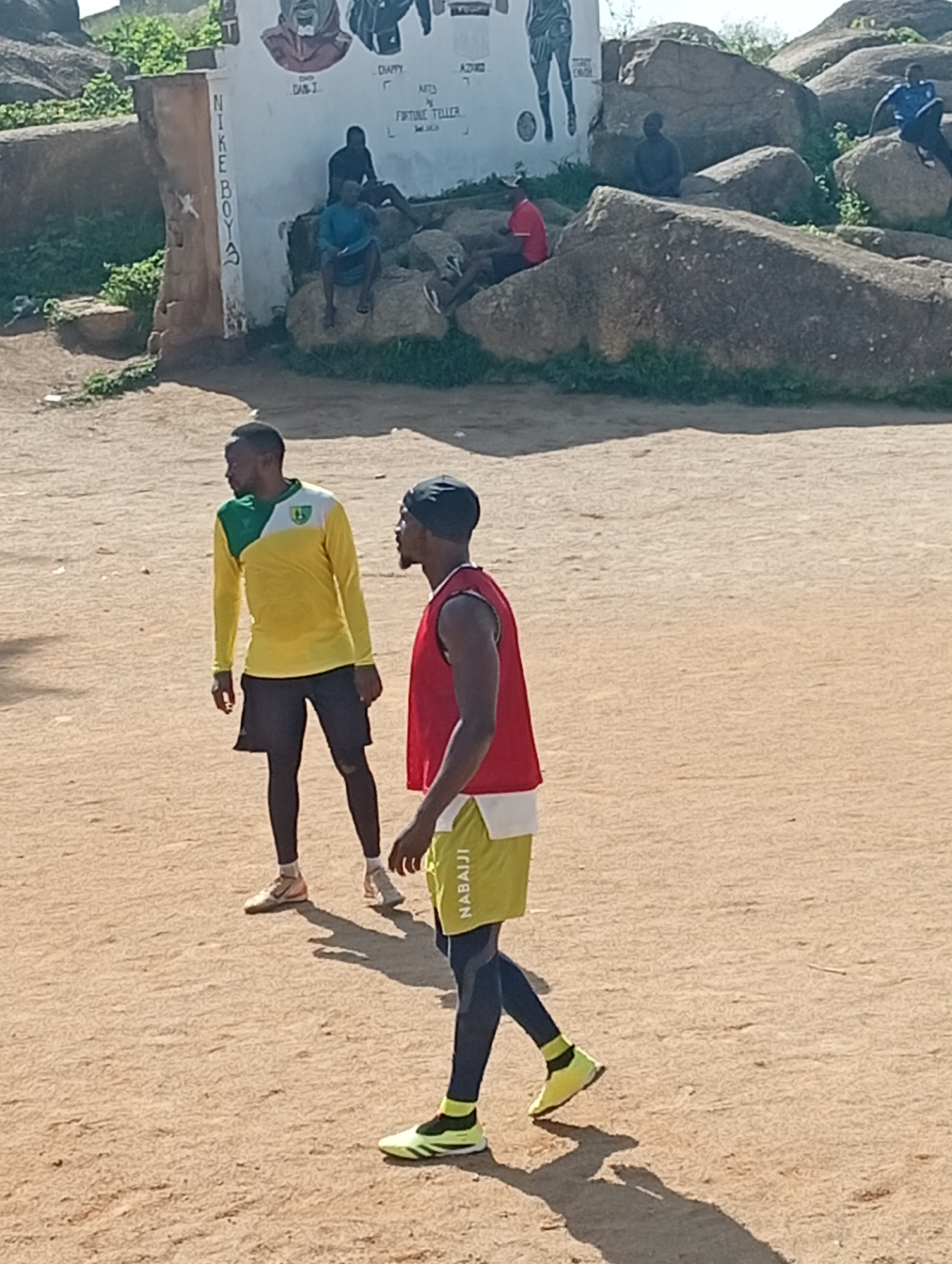 Two young men are playing soccer in an open field in a rocky, arid environment, possibly in an African city. In the background, there is a wall with graffiti and some large rocks. One man, wearing a yellow and green long-sleeved shirt and black shorts, is standing and looking to the right. The other man, wearing a red tank top and yellow shorts with black leggings, is also standing and looking to the right. Both men are wearing athletic shoes. There are other people in the background, some sitting on rocks and others standing. The ground is a light brown, sandy dirt.