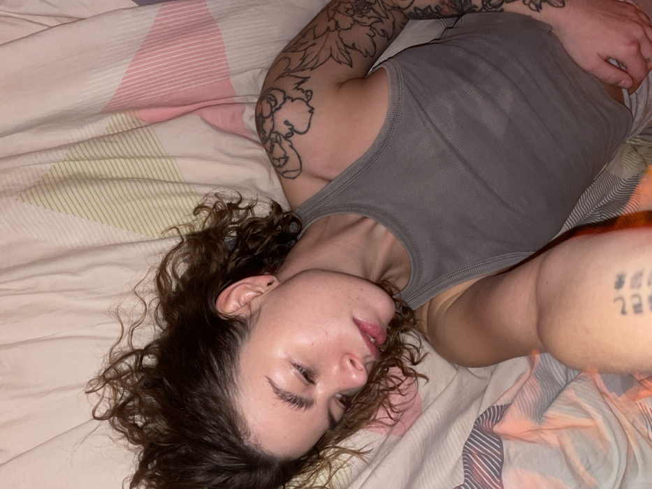 A young woman with curly brown hair lies in bed on striped sheets. She wears a gray tank top and has tattoos on both arms. Her eyes are closed, and her lips are slightly parted. The lighting is soft, and her skin appears dewy.