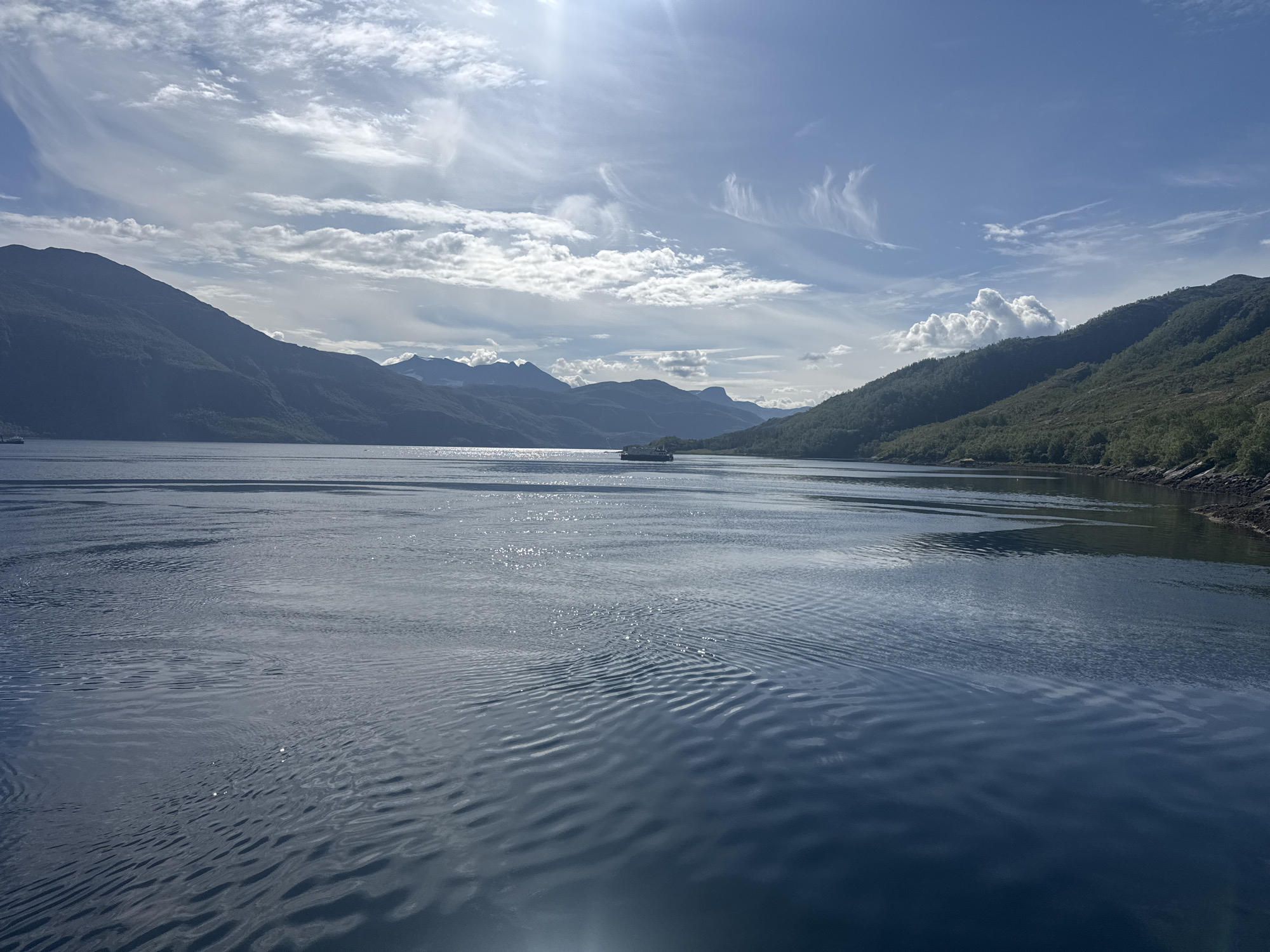 The image shows a wide, calm body of water, likely a fjord, with steep, tree-covered mountains on either side. The sky is blue with scattered white clouds, and the sun is shining, creating reflections on the water. In the distance, a small ferry boat is visible. The overall impression is one of a serene and beautiful natural landscape. The image was taken from the water, looking towards the shore. The location is Inndyr, Norway.