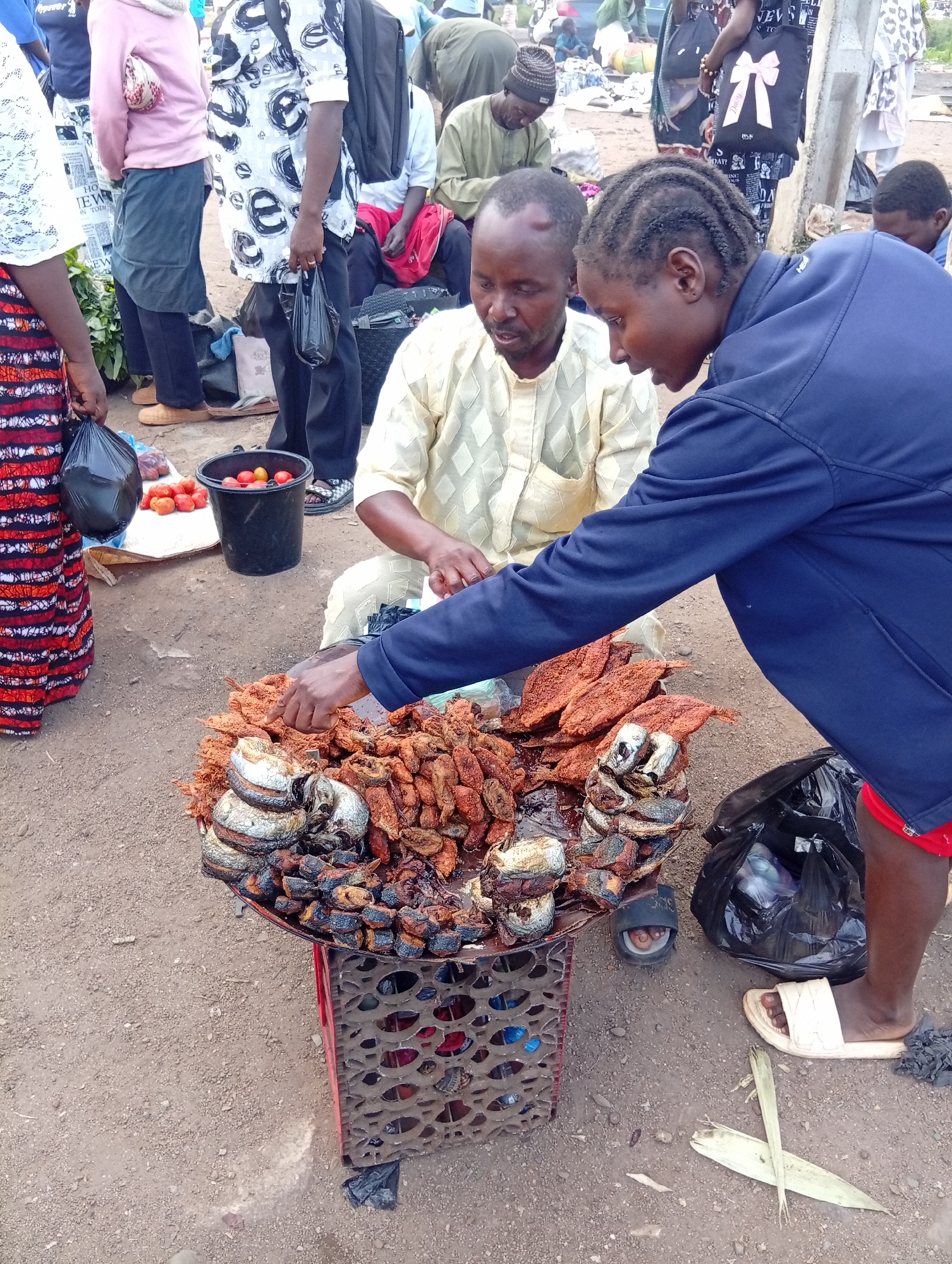 In Bukuru, Nigeria, a street vendor displays fried fish and tomatoes. The vendor, a man wearing a light yellow embroidered traditional outfit, stands behind a cart laden with fried fish. A woman, with her hair in braids and wearing a dark blue jacket, crouches to interact with the vendor. She is pointing at the fish, possibly discussing a purchase. In the background, other individuals and market stalls are visible, suggesting a bustling marketplace environment. A black bucket filled with red tomatoes sits on the ground near the vendor. The ground is dirt and gravel.